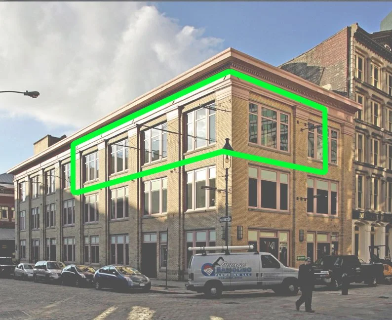 A three-story corner brick building with large windows outlined in green, located on a city street with parked cars and a person walking past.