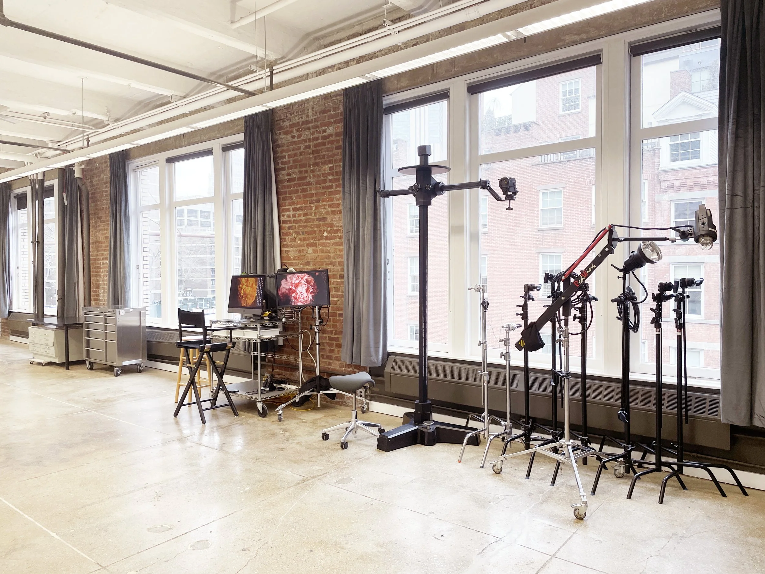 A bright indoor space with large windows, curtains, and various equipment including tripods, monitors, and desk items, suggesting a studio or workspace.