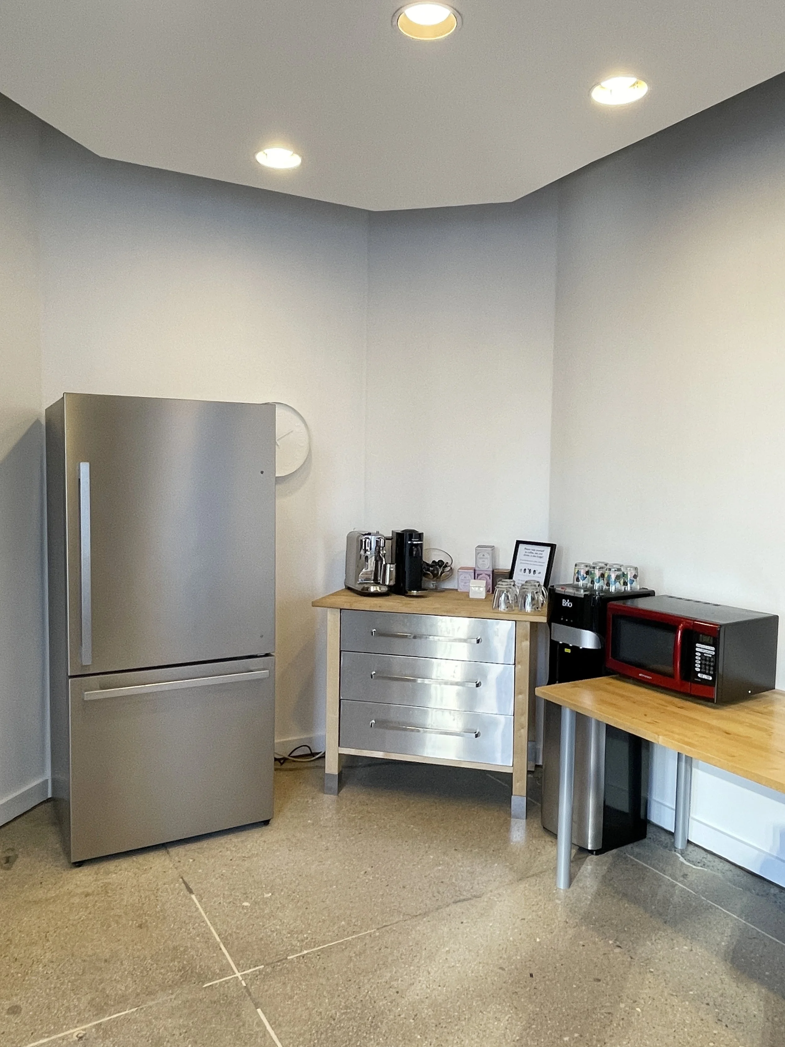 A small kitchenette area with a stainless steel refrigerator on the left, a wooden counter with a coffee maker and cups in the middle, and a black and red microwave on the right, all on gray tables against gray walls with a white ceiling and recessed lighting.