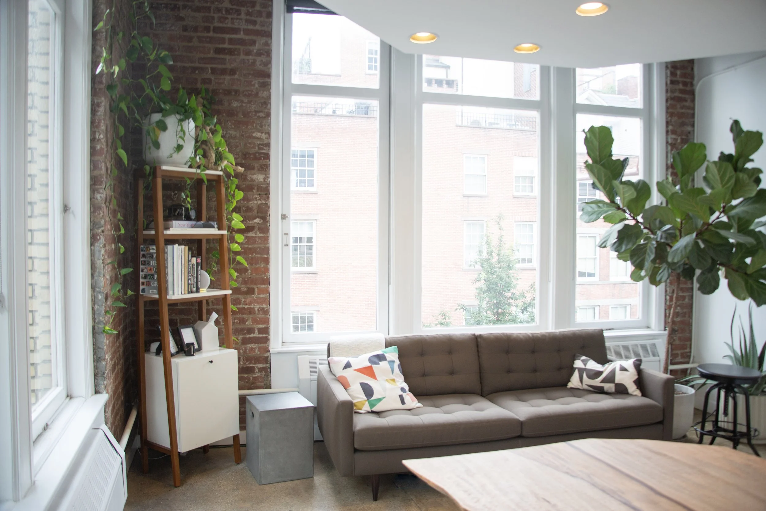 A cozy living room with large windows revealing brick buildings outside. It has a modern sofa with colorful geometric pillows, a wooden bookshelf with plants and books, and a mix of exposed brick and white painted walls. Recessed ceiling lights illuminate the space.