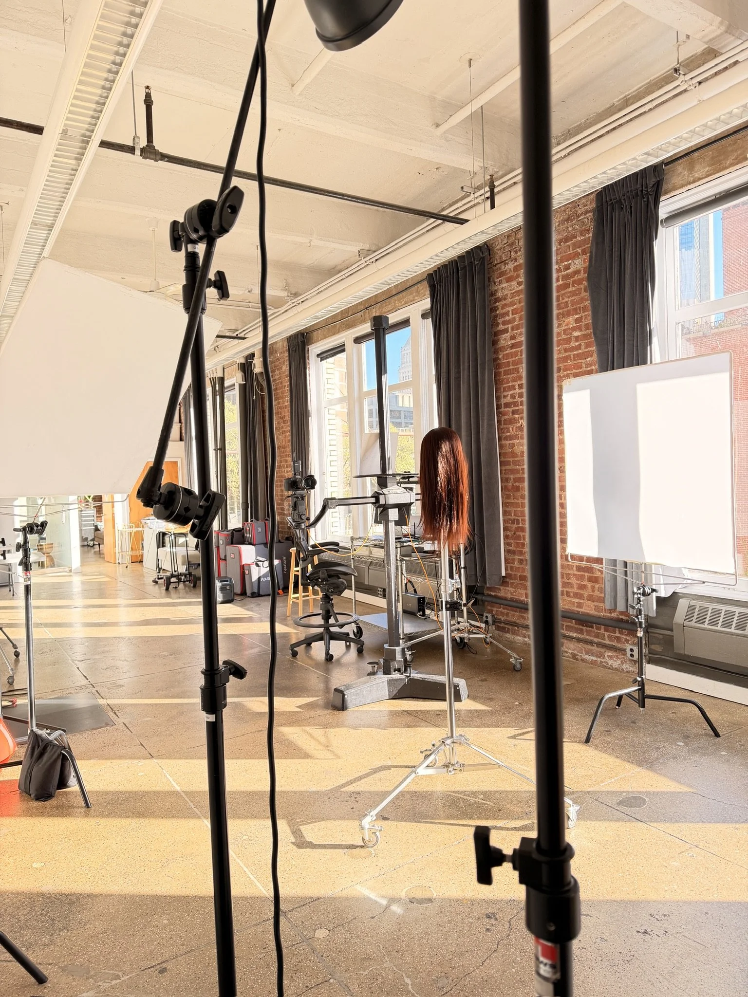 Interior view of a photography or video studio with equipment such as lighting stands, a camera, and a mannequin with long red hair, set against large windows and brick walls.