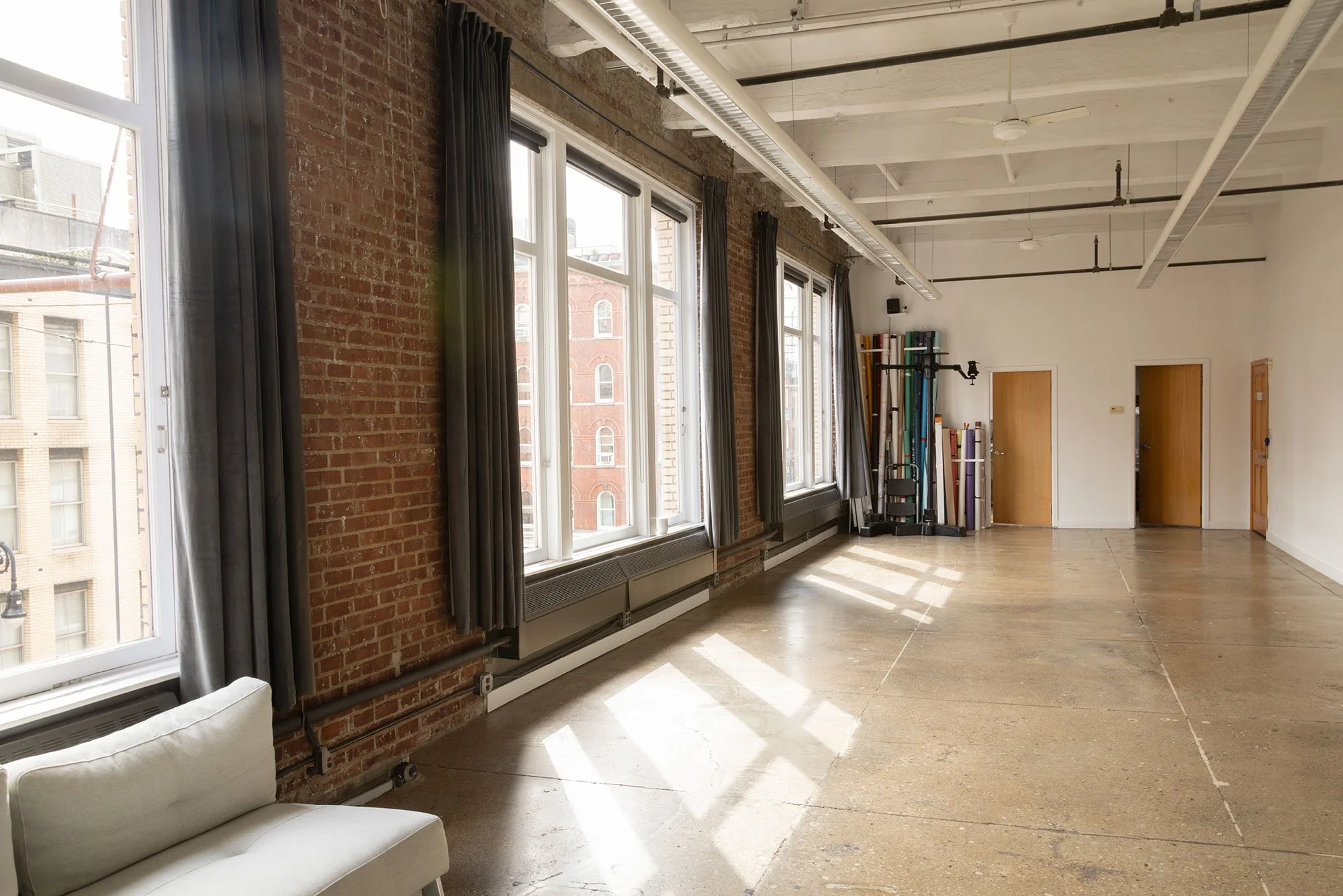A large room with large windows, exposed brick wall, wooden floor, and various equipment and materials stored along the back wall. Sunlight is streaming through the windows.