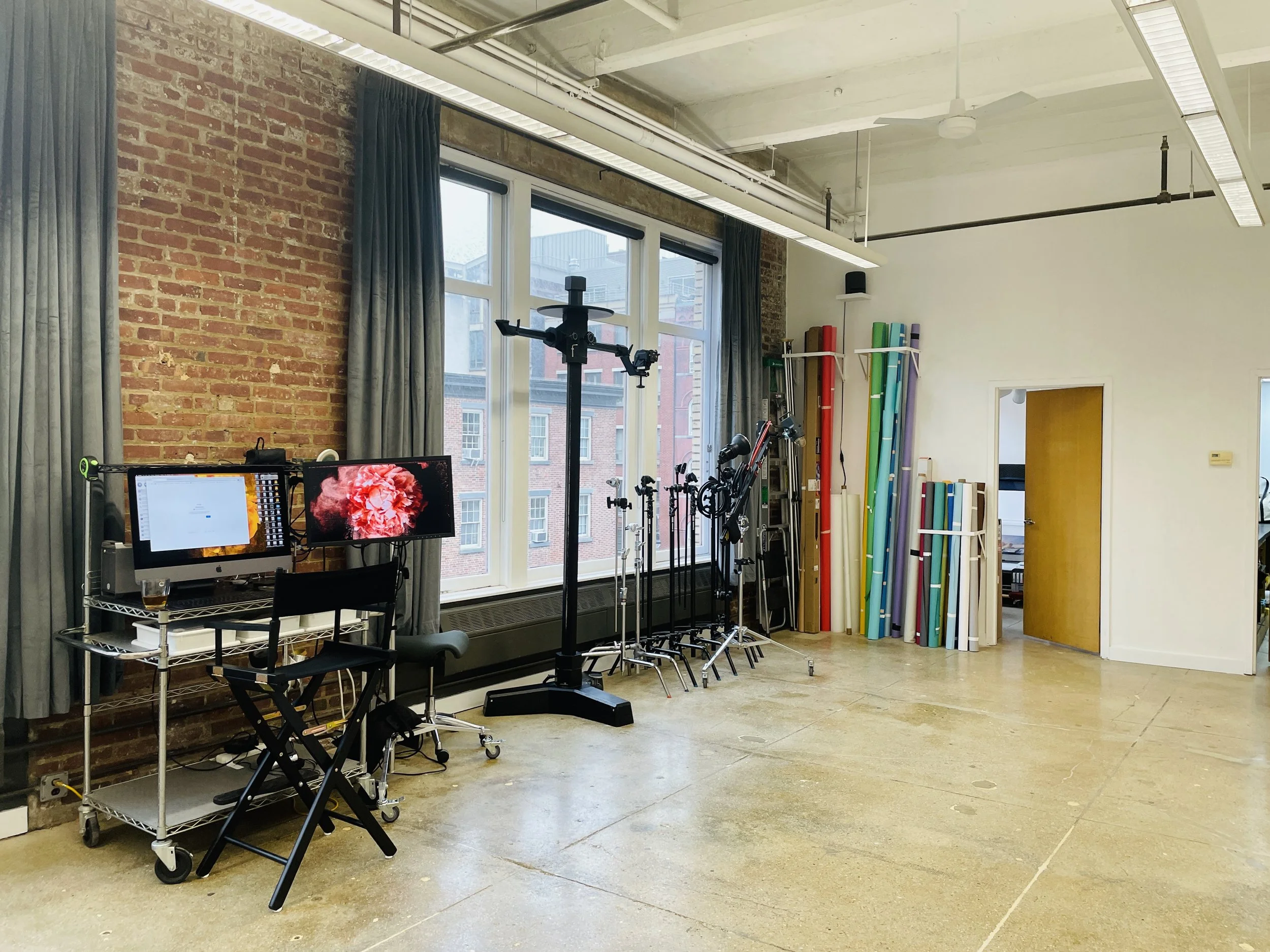 An indoor studio or workspace with large windows, brick walls, and various equipment including monitors, tripods, and colorful rolled mats or tubes, with a permission for natural daylight.