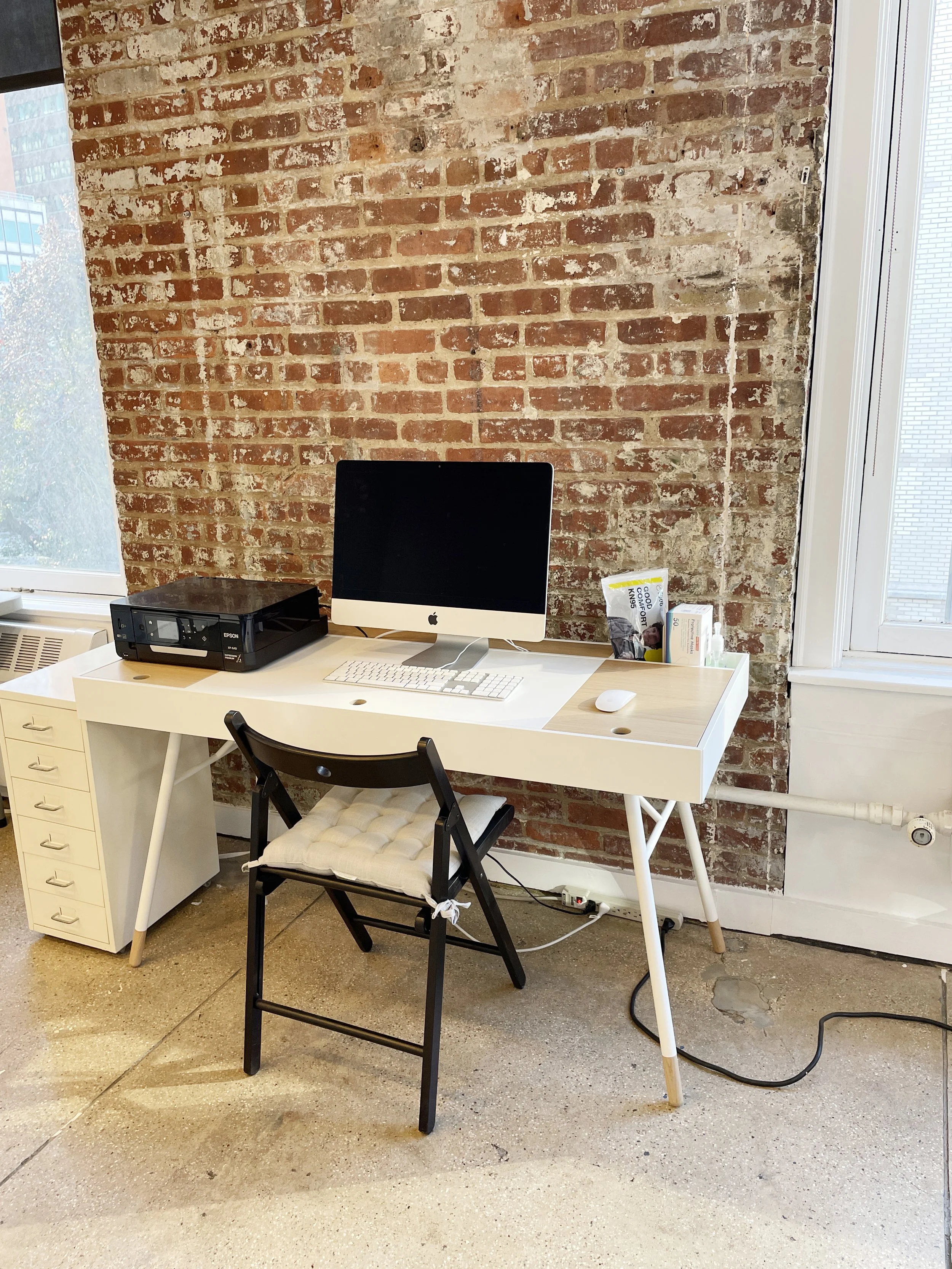 An office workspace with a white desk, a computer, a printer, and office supplies against a brick wall. There is a black chair with a cushioned seat and a window to the right.