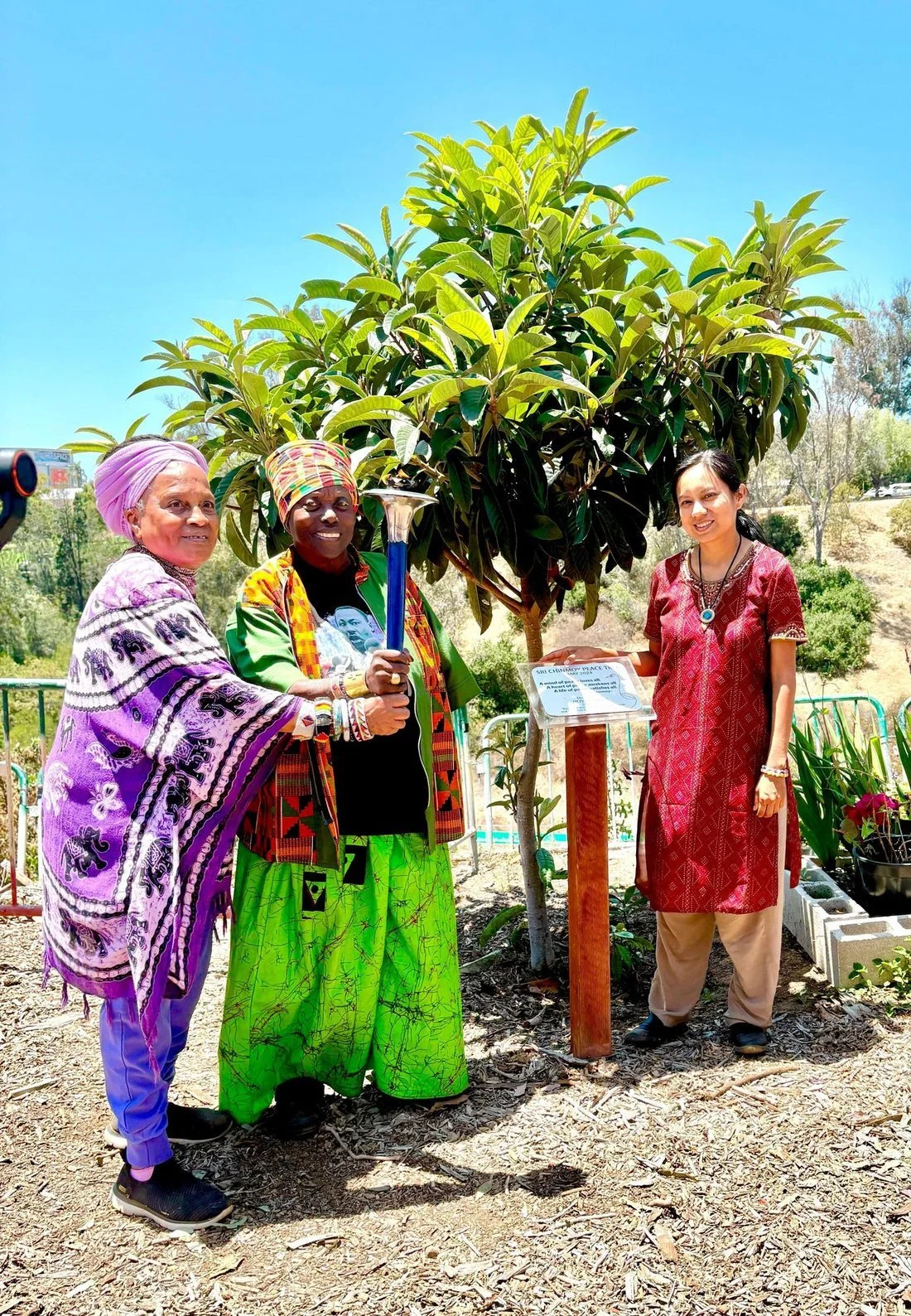 Three women standing outdoors in front of a large tree, one holding a ceremonial torch, with a sign on a stand beside them.