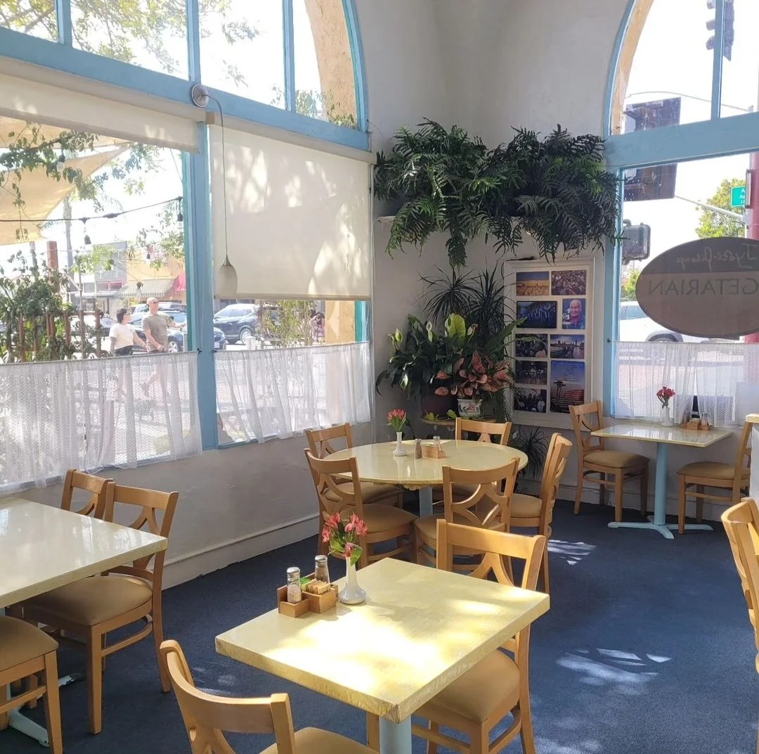 Interior of a cozy restaurant with light-colored tables and wooden chairs, decorated with small vases of pink flowers, surrounded by large windows and potted plants in the corner.
