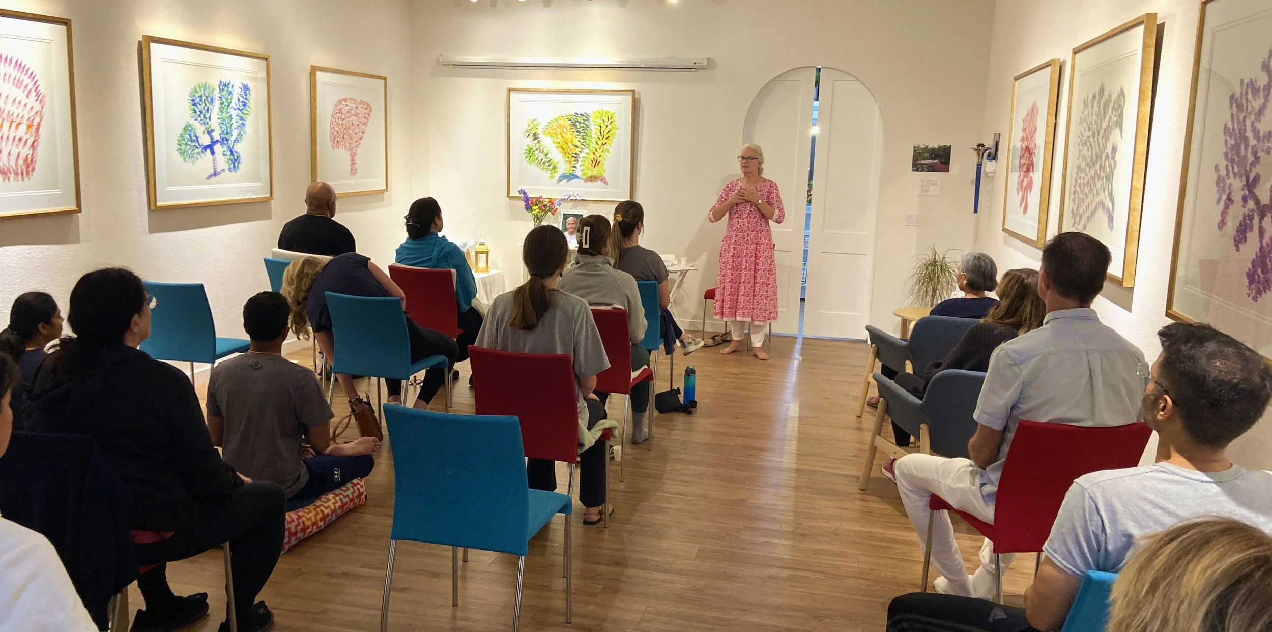 Woman in pink dress leading a presentation in art gallery with audience seated in colorful chairs, framed artwork on walls.