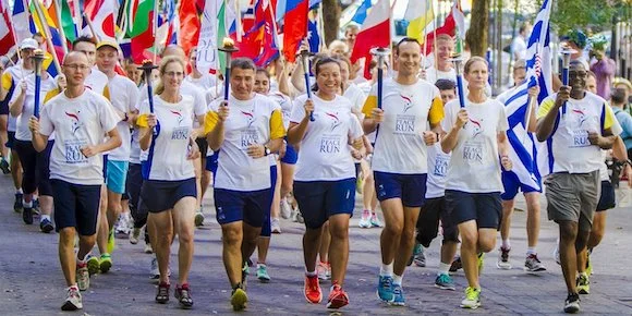 Group of people participating in a fun run or parade, running on a street with flags in the background, wearing matching white T-shirts with a logo.