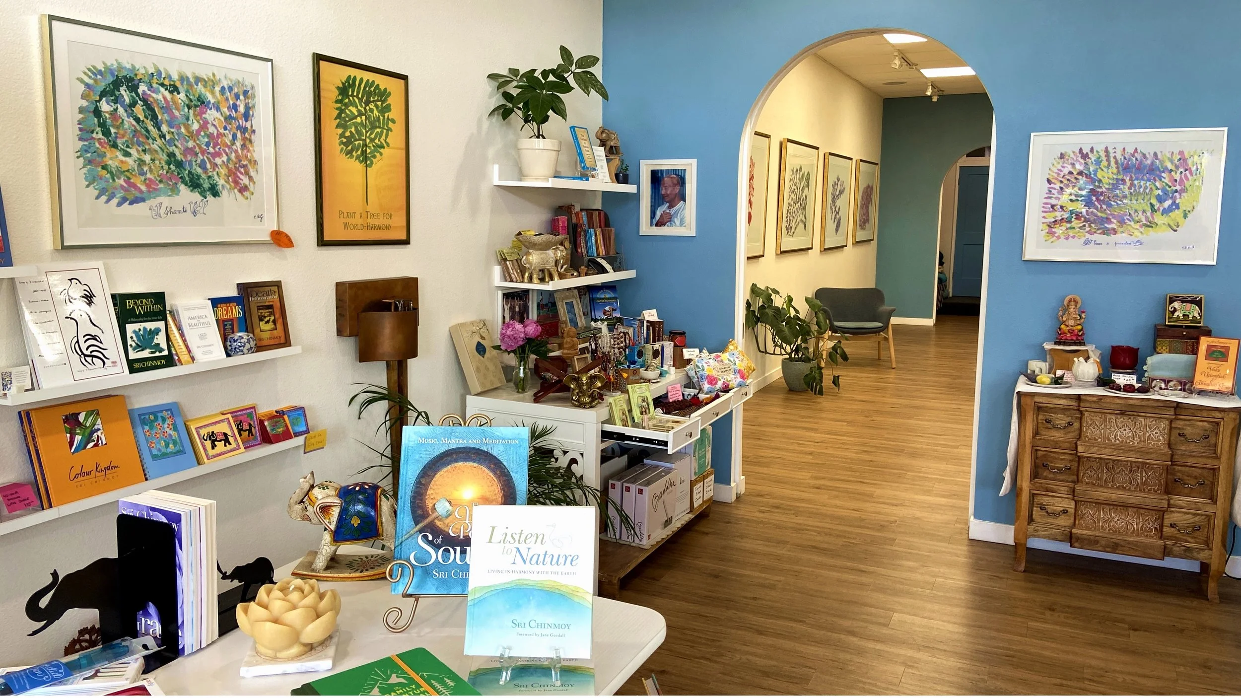 Interior of a bookstore or gift shop with various shelves and framed artwork on the walls. The space features brightly colored books, decorative items, and plants, with a wooden floor and a blue accent wall.
