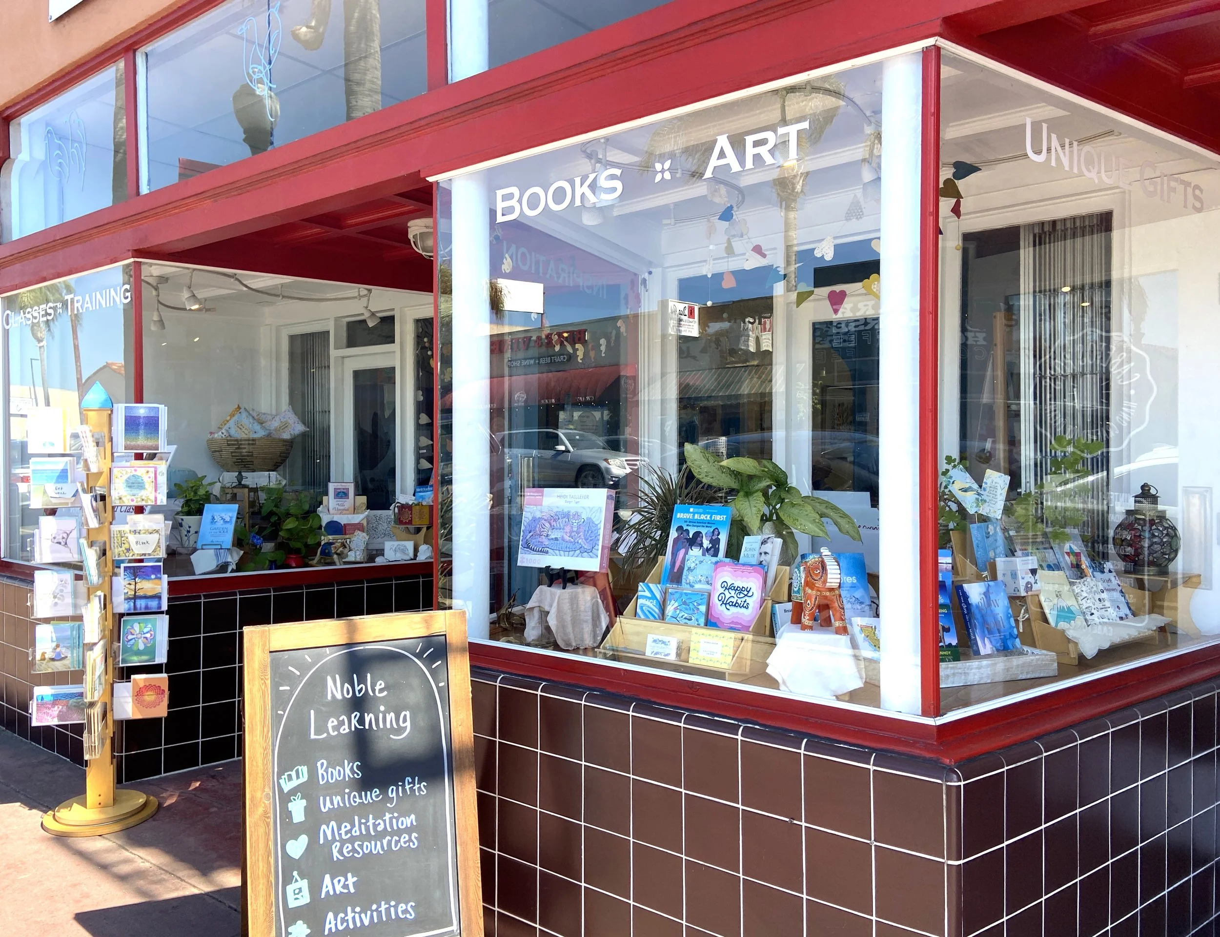 A shop window with a sign advertising books, art, and unique gifts, with a chalkboard sign outside listing the shop's offerings including classes, learning, meditation resources, art, and activities. The shop has a red frame and large glass windows showcasing books and decorative items inside.