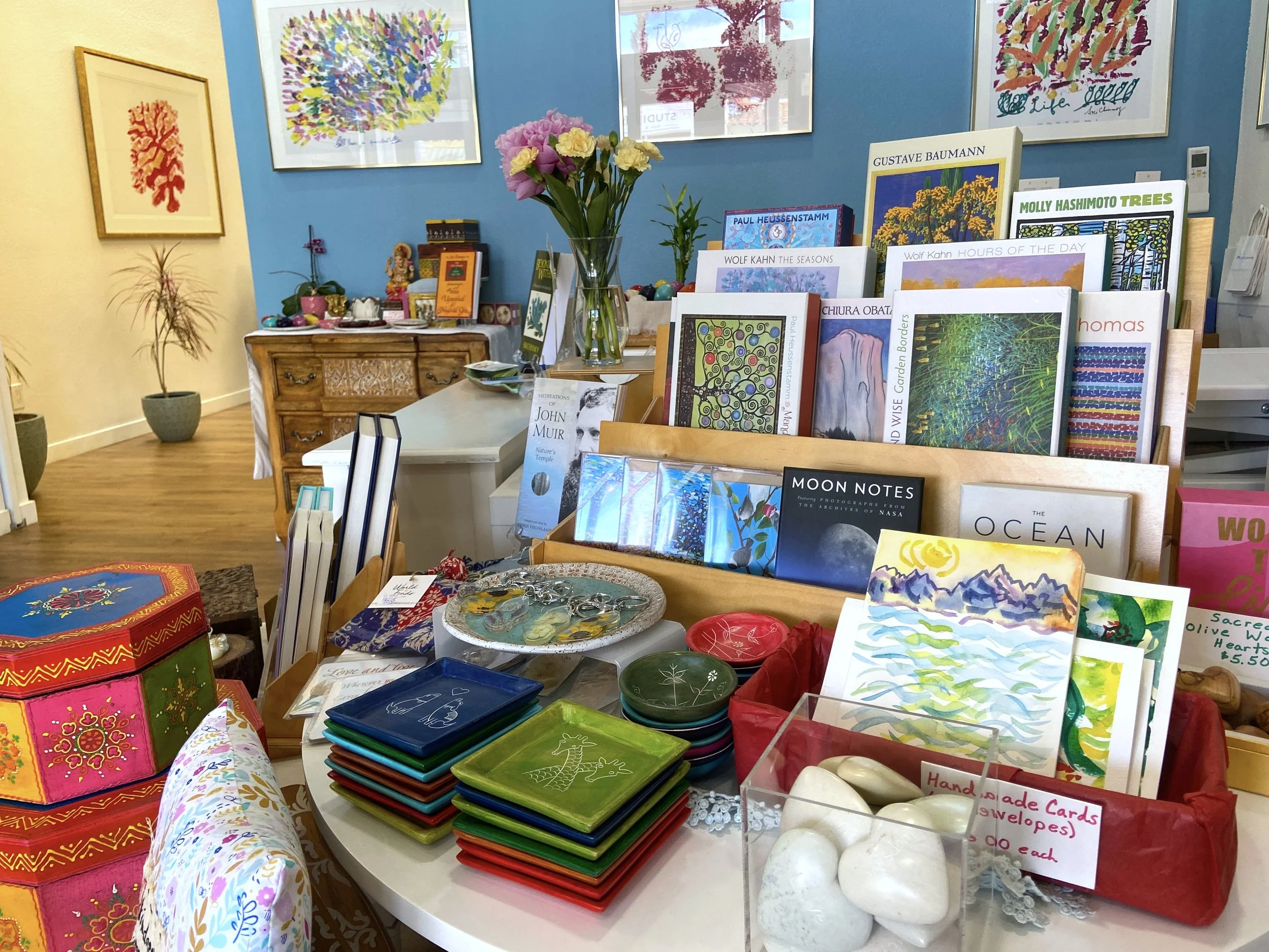Inside a gift shop with colorful greeting cards, decorative bowls, and framed artwork on the walls. A vase with pink and yellow flowers is on a table, and various books and crafts are displayed on tables and shelves.