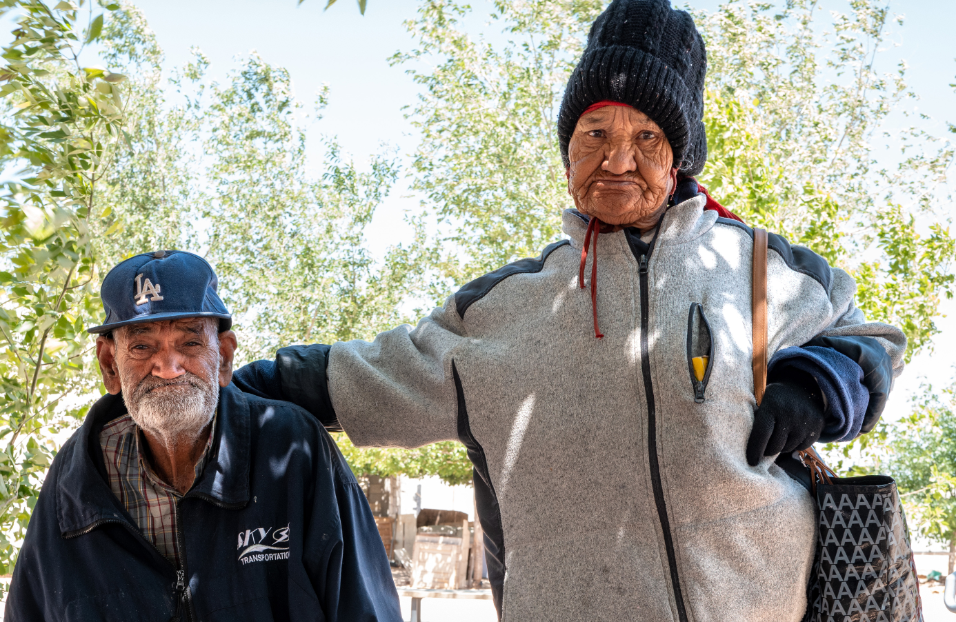 Two elderly people outdoors at Punto Zero, one man with a gray beard and a blue baseball cap with LA on it, and a woman wearing a black beanie and gray zip-up jacket, with trees and a sunny sky in the background.