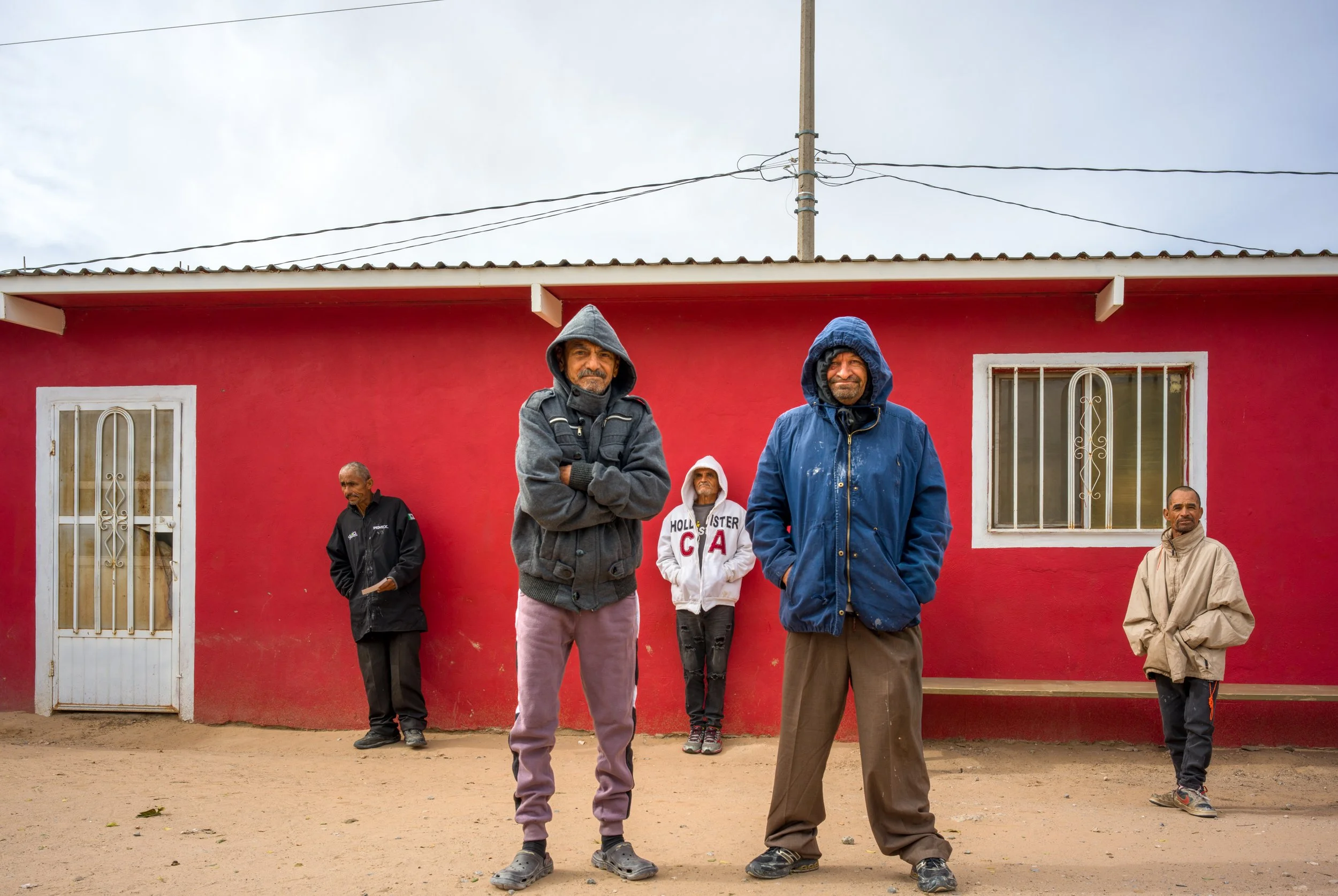 Group of six men standing outside in front of a red building with barred windows, some wearing hooded jackets, with overcast sky above.