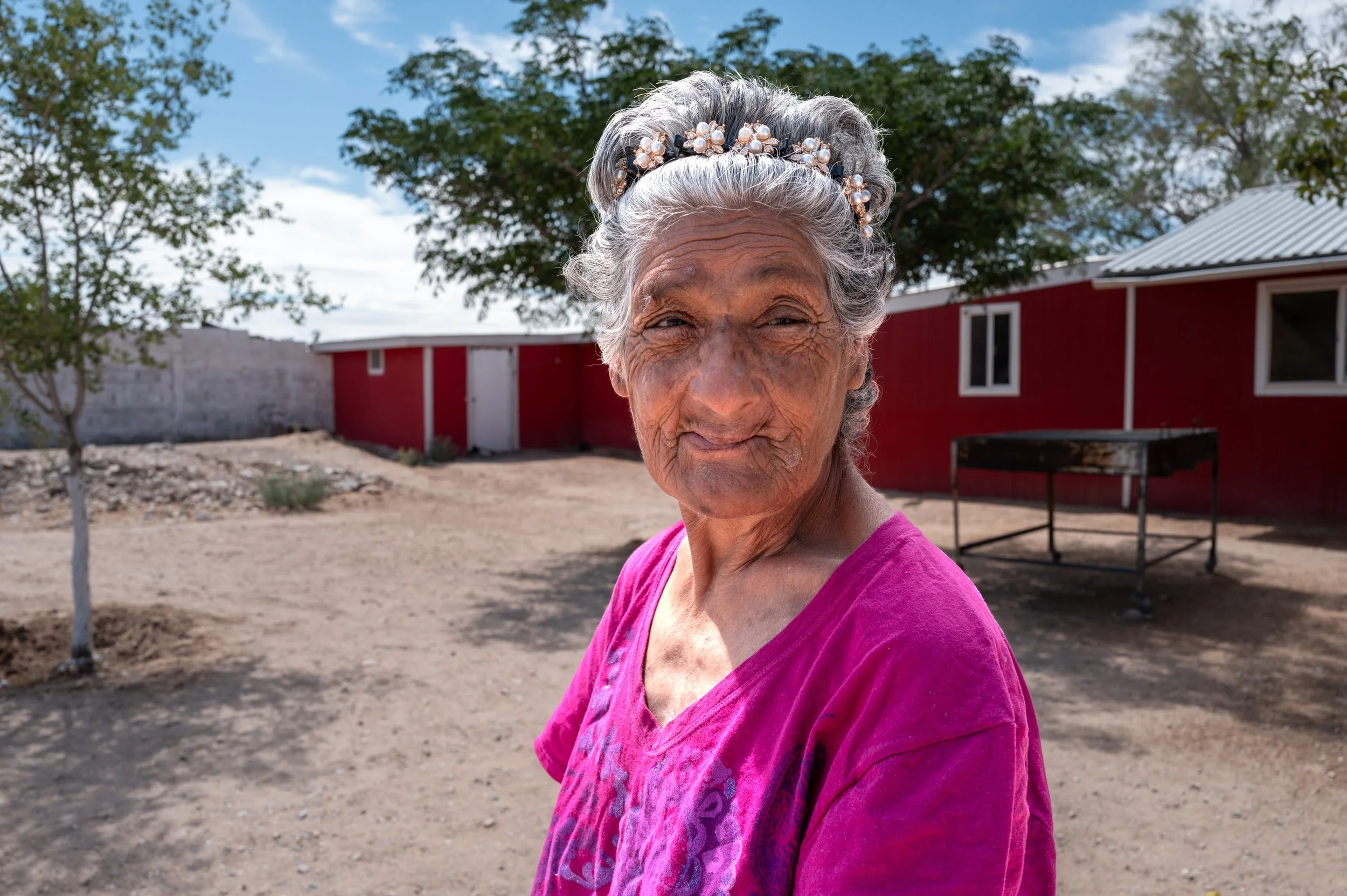 An elderly woman with gray hair styled in an updo at Punto Zero, wearing a floral headband and a pink shirt, standing outdoors with trees and a red building in the background.