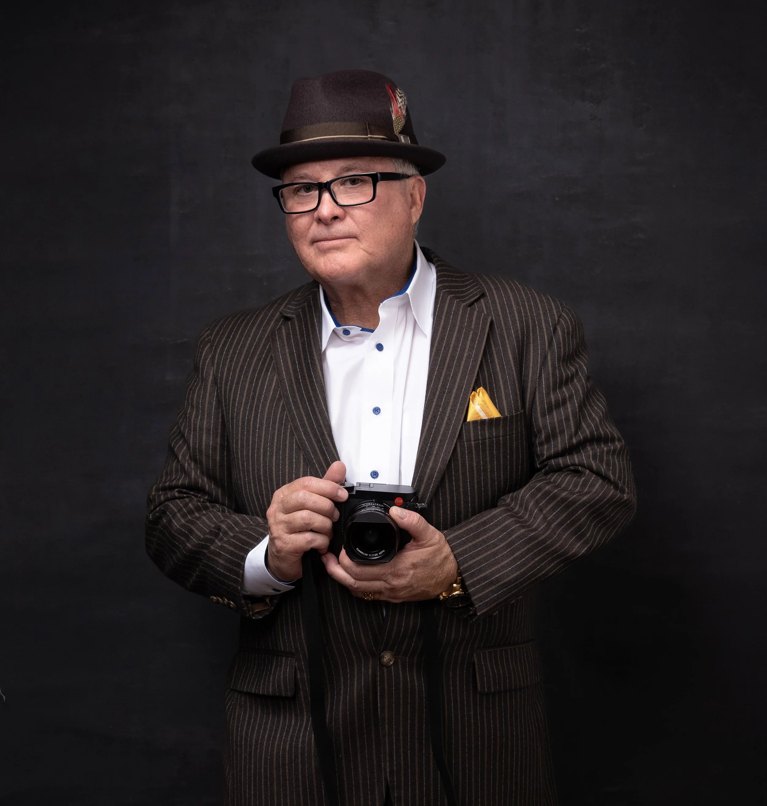 A man wearing glasses, a striped suit, and a fedora hat with a feather, holding a camera in front of him, standing against a dark background. Art Moreno Jr., author of Dignity Has A Name.