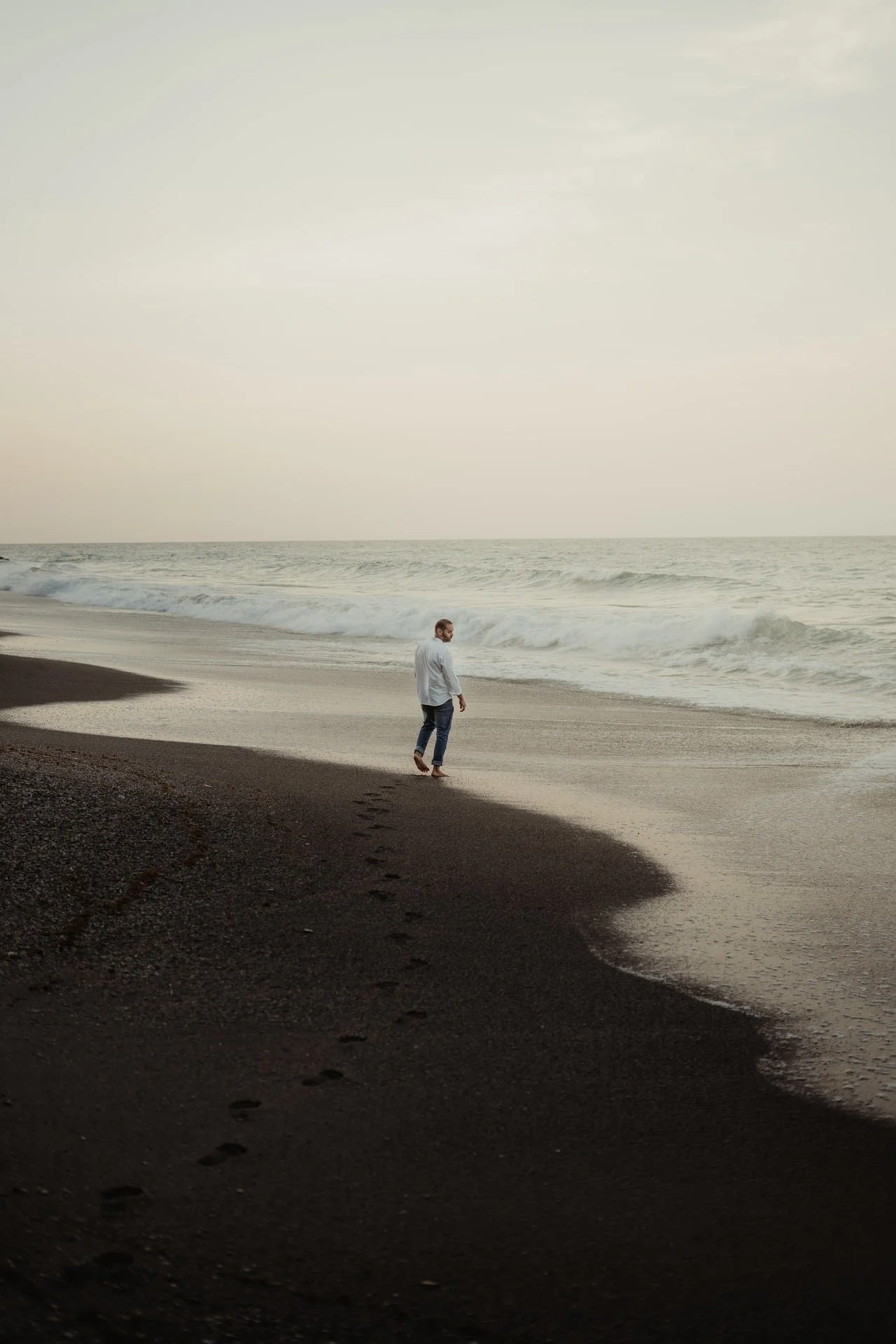 A person walking along the sandy shore of a beach, with waves in the background under a cloudy sky.