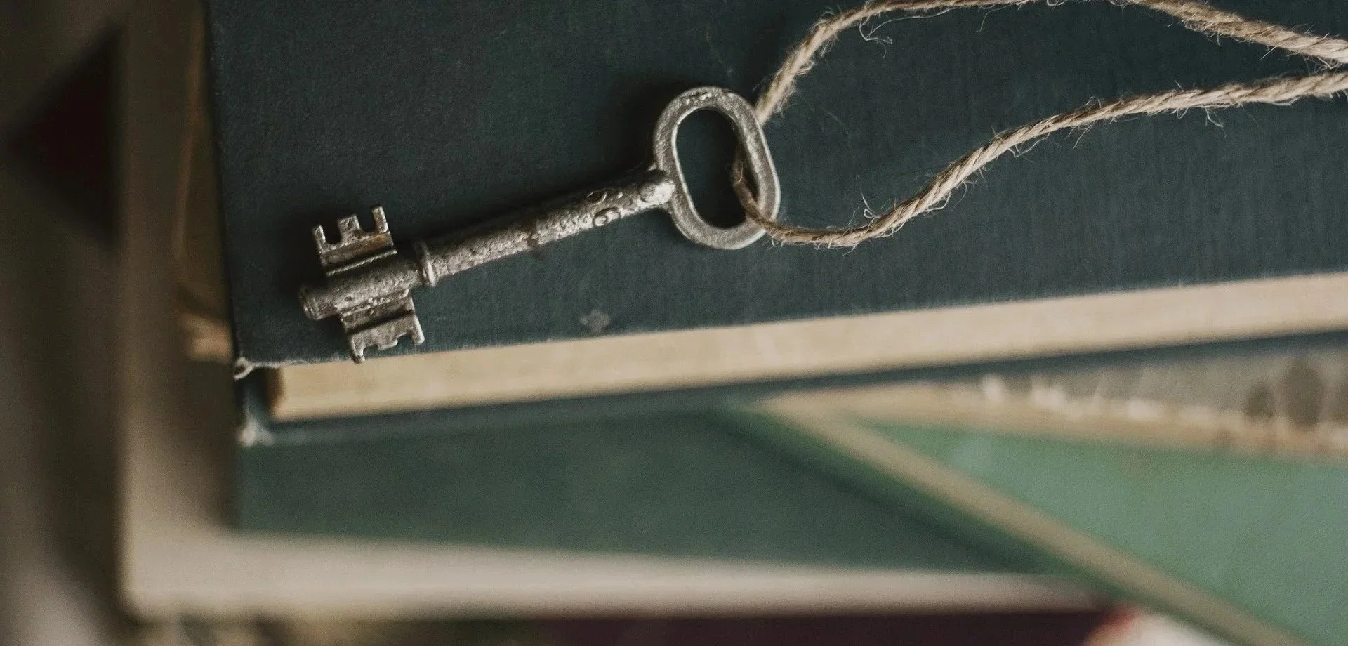 An antique metal key with a rustic finish hanging from a piece of twine. The key is placed on top of a dark-colored book or box.