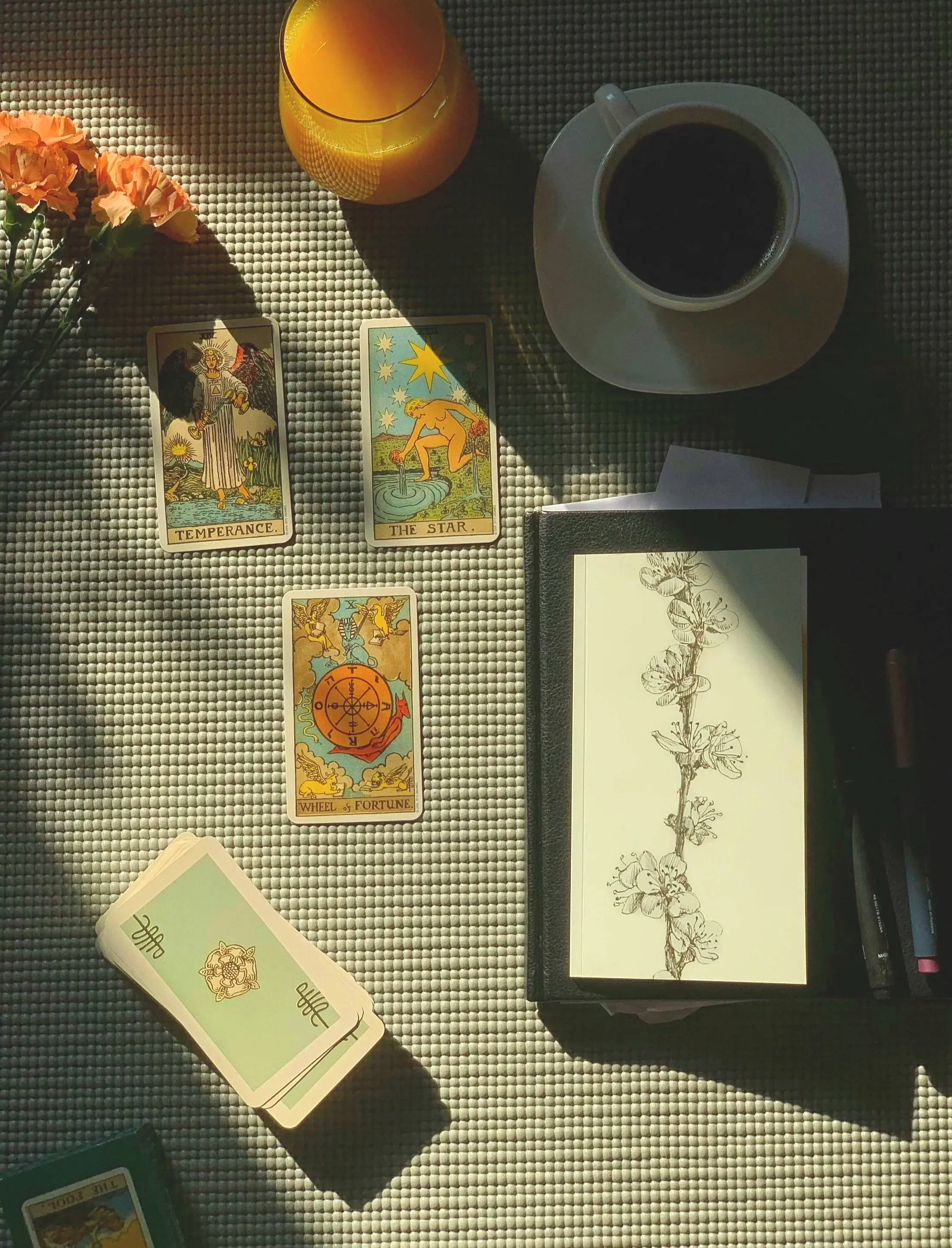 Tarot cards spread out on a textured surface, including the Temperance, The Star, and Wheel of Fortune cards, with a cup of black coffee, a glass of orange juice, a bouquet of orange flowers, and a black notebook with botanical sketches nearby.