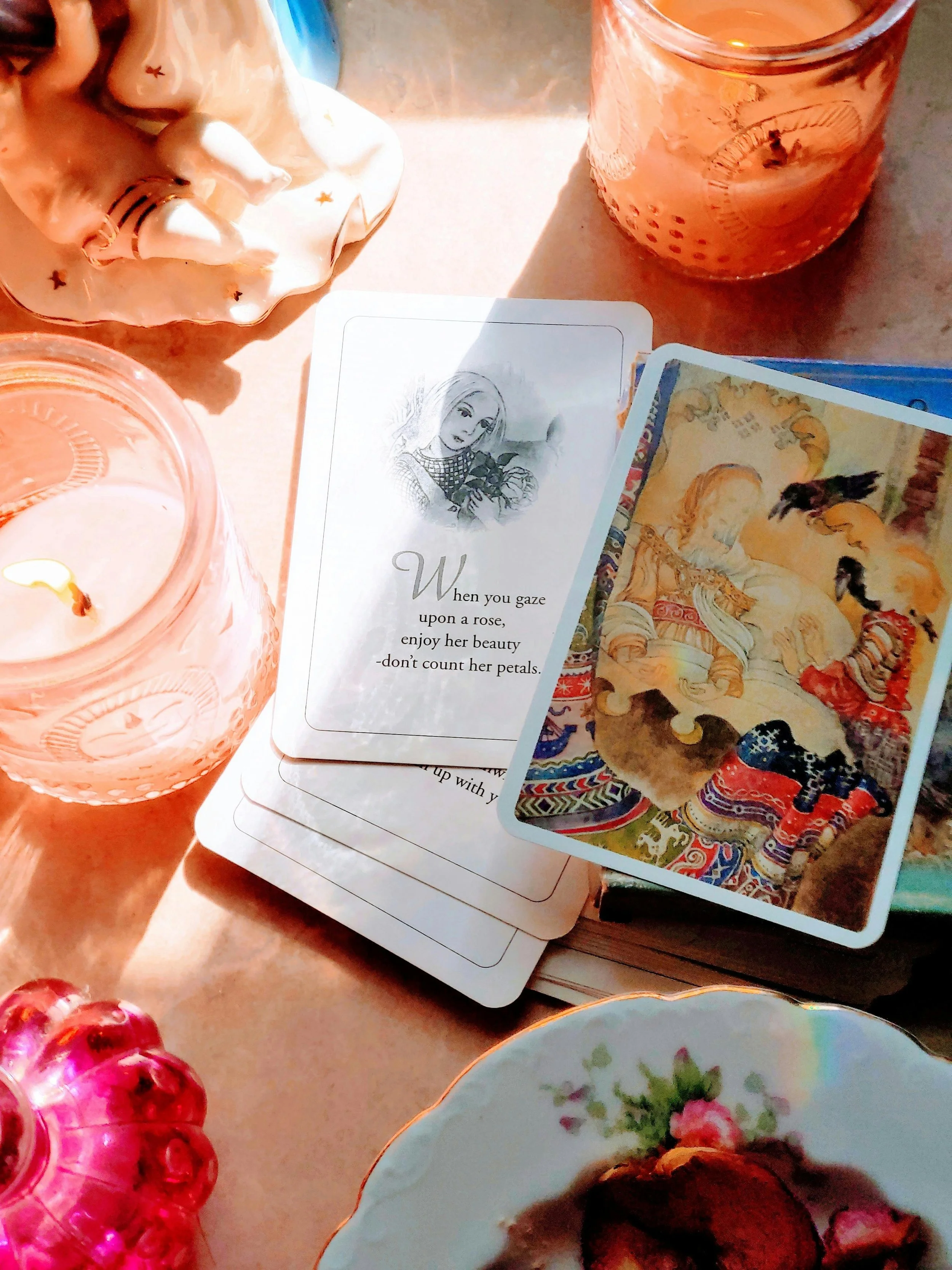 A collection of decorative items on a table, including a candle in a pink glass holder, a deck of tarot or oracle cards featuring artwork of a woman with long hair, and a porcelain bowl with a floral pattern containing a baked good. There are also two glasses, one pink and one clear, with textured designs.