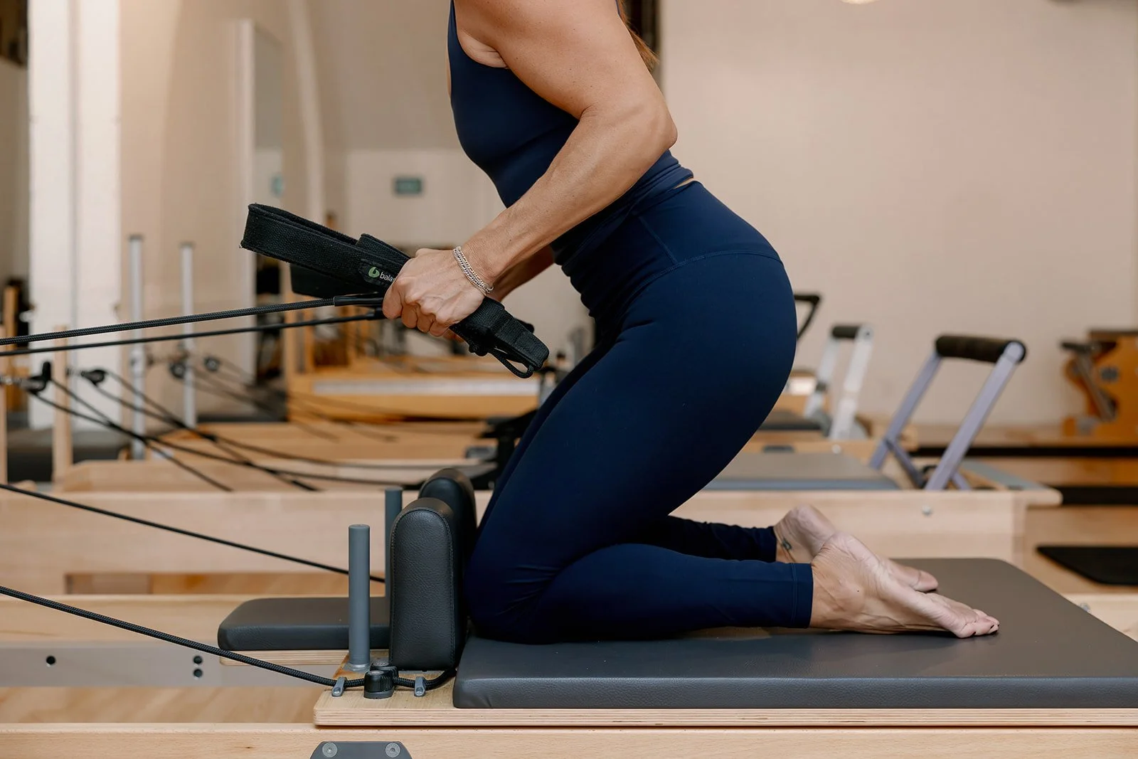 A woman kneeling on a Pilates reformer machine, pulling on resistance bands, in a fitness studio.