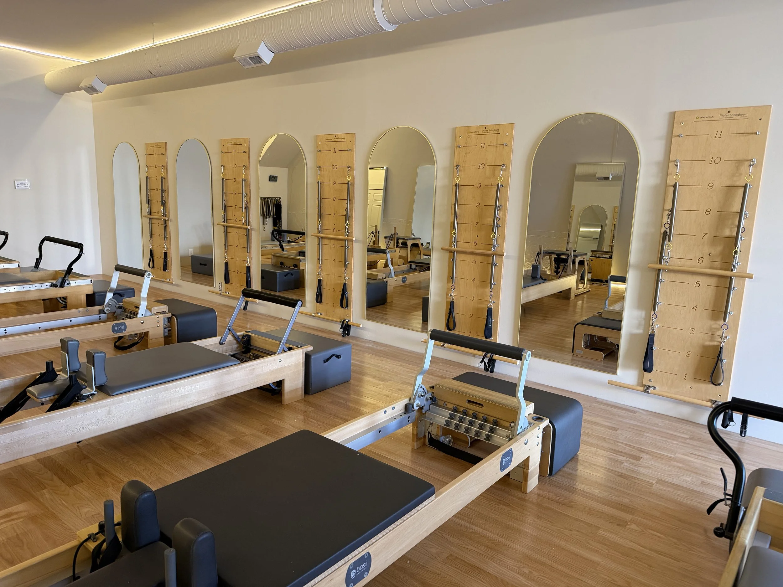 A Pilates studio with several wooden reformer machines and wall-mounted balance boards with measurement lines and straps, mirrored wall, wooden flooring, and ceiling vents.
