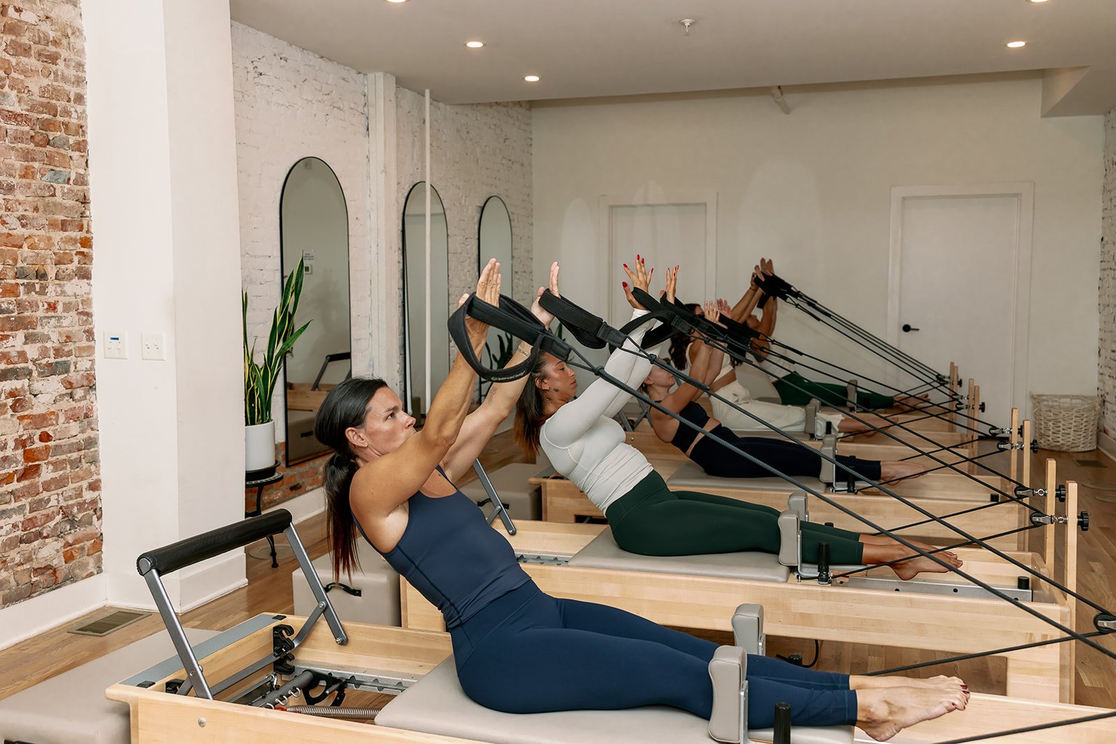 A group of women participating in a Pilates reformer class, exercising with reformer machines in a bright studio with white walls, mirrors, and wooden floors.