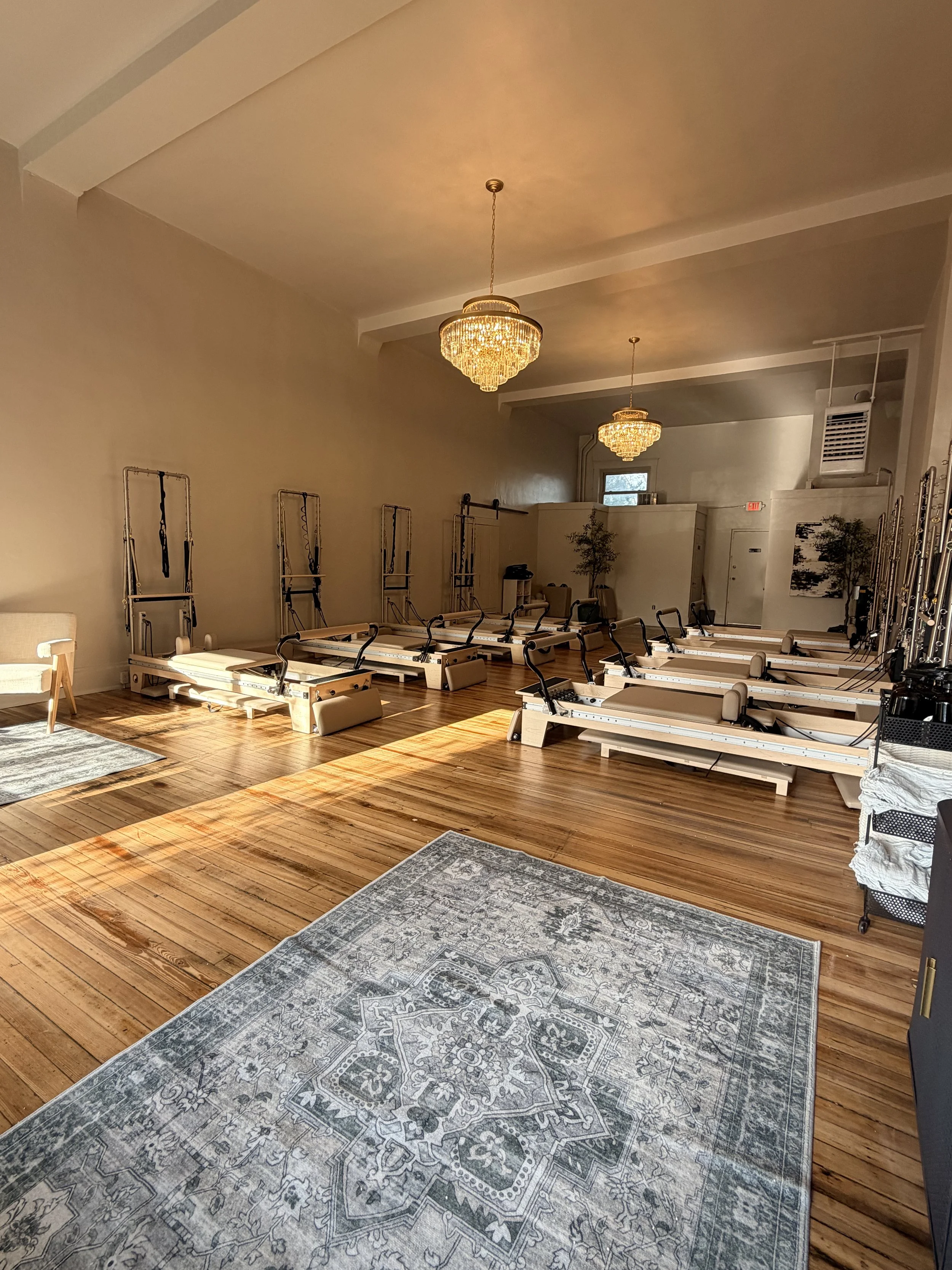 A spacious yoga or Pilates studio with wood floors, exercise reformers lined up against the walls, chandeliers hanging from the ceiling, and area rugs on the floor.