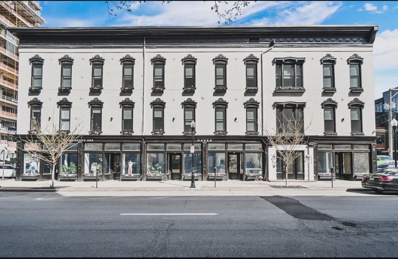 A four-story white brick building with black window and door frames, situated along a city street with leafless trees and parked cars.