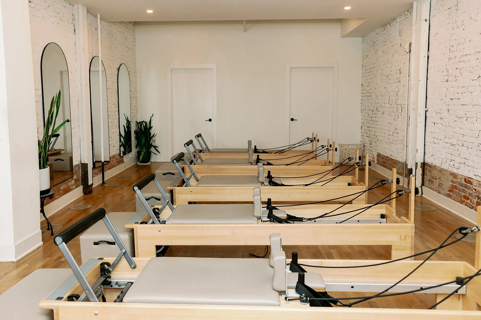 A Pilates studio with six reformer machines, potted plants, three mirrors on the wall, and white brick walls.