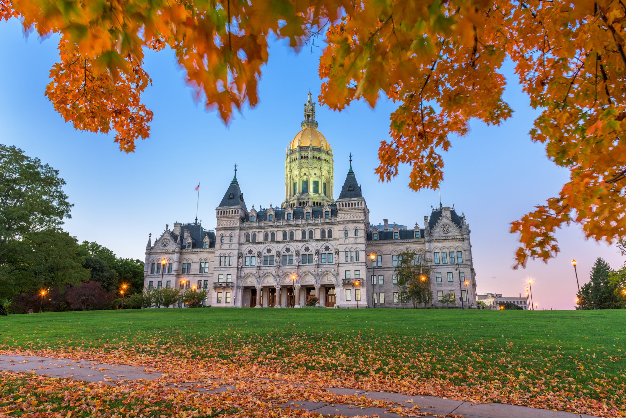 Autumn leaves in orange and yellow surround a historic building with a golden dome and turrets, situated in a park with a well-kept lawn and trees, under a clear blue sky.