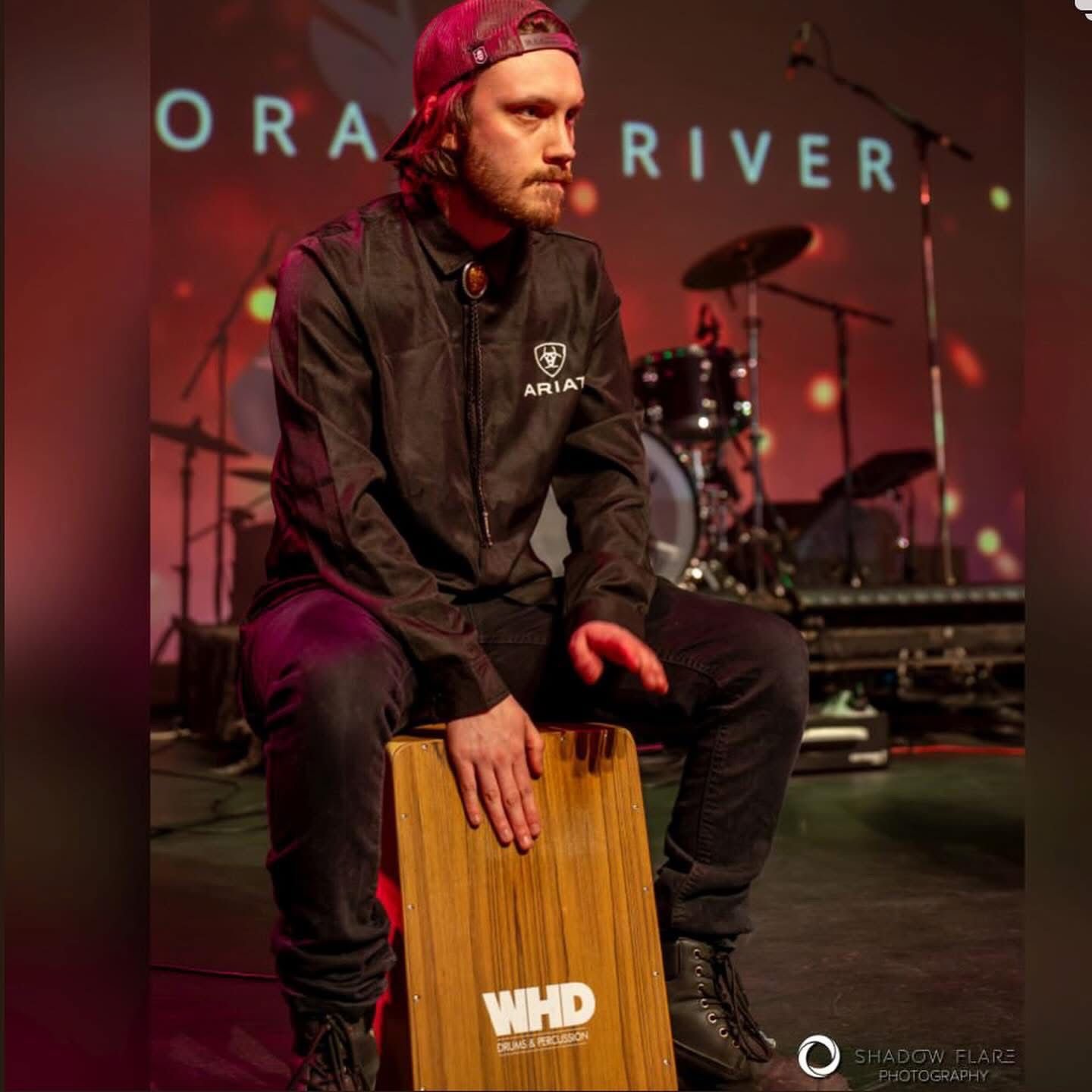 A man sitting on a cajón on stage, wearing a black ARIAT jacket and a red cap backwards, with a drum set and stage lighting in the background.