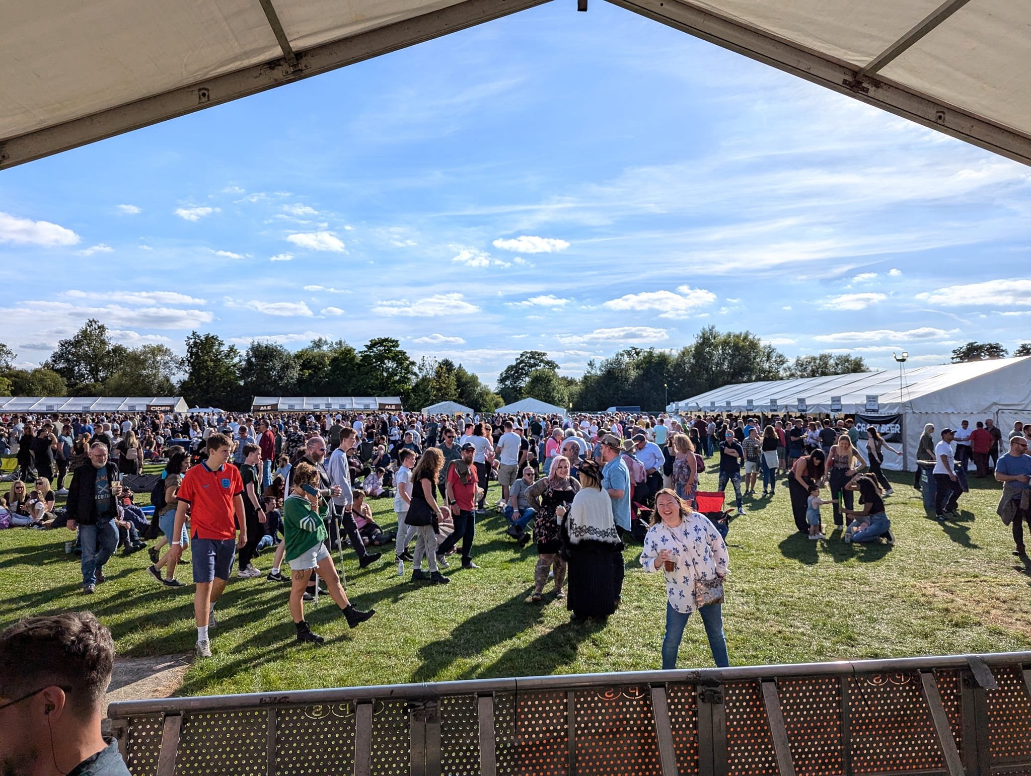 A crowded outdoor festival with people walking and standing on green grass, tents in the background, and a blue sky with clouds overhead.