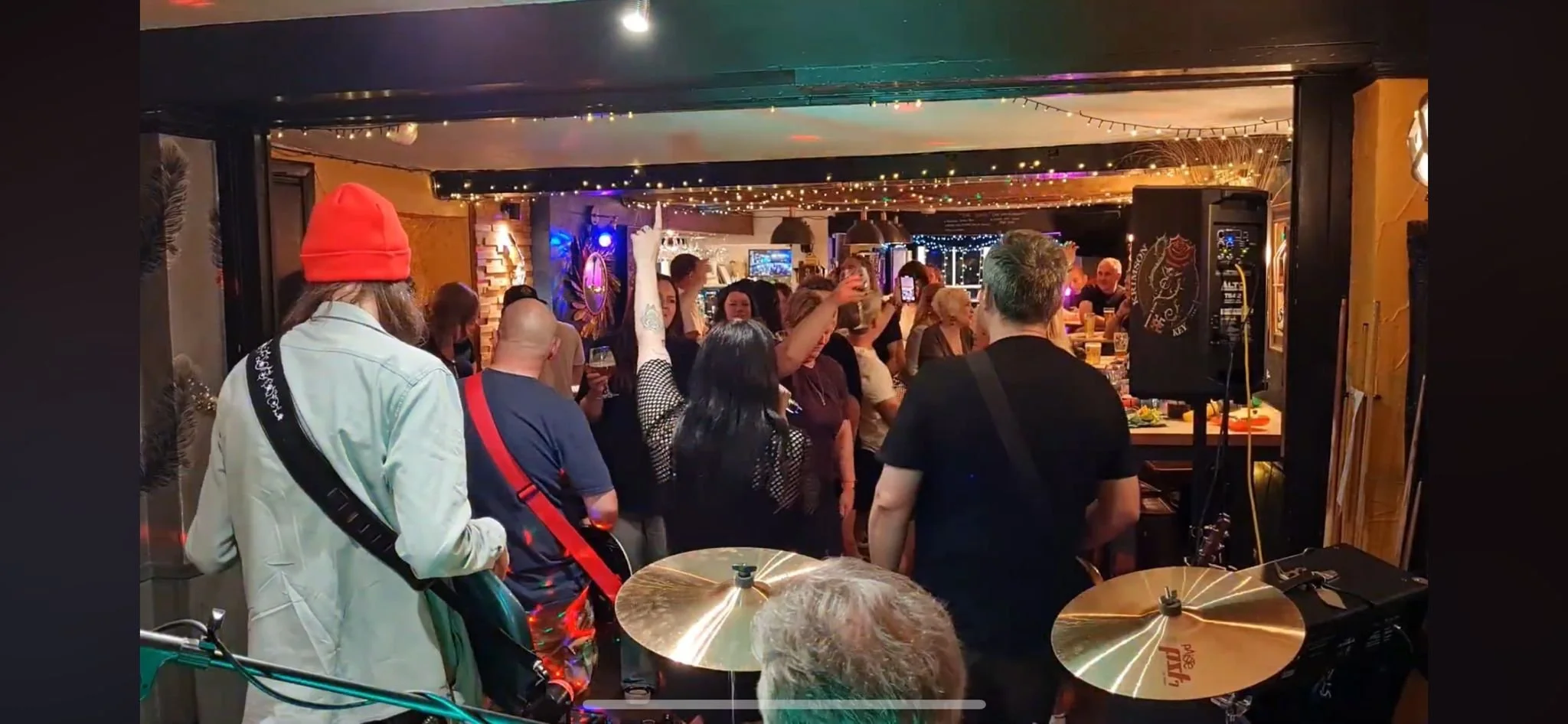 A group of people enjoying a night at a bar or pub with colorful string lights on the ceiling. Some are holding drinks, and one person is raising their hand. There is musical equipment, including cymbals, in the foreground, suggesting live music.