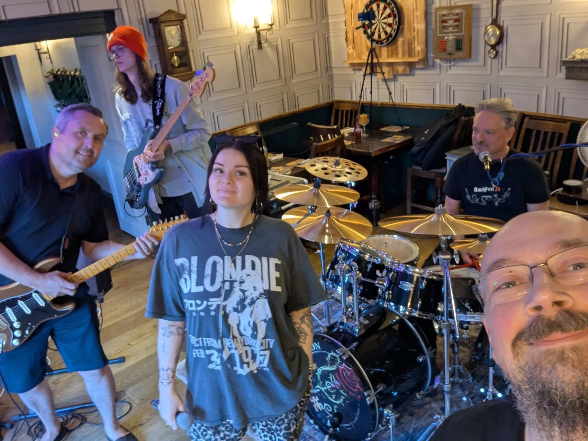 A group of five people in a cozy, wood-paneled room with musical instruments and a dartboard. Two are seated near drums, one stands with a bass guitar, and two are on the right side, one with headphones. The atmosphere suggests a casual band practice or gathering.