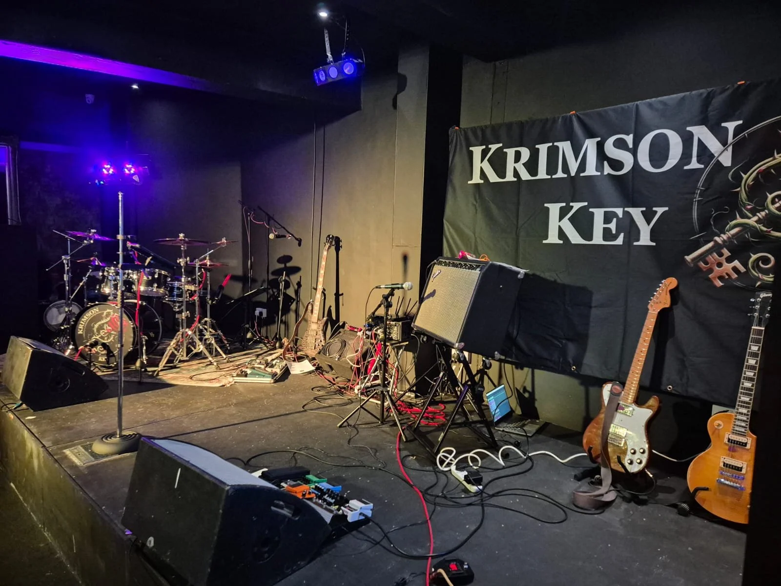 Concert stage with drums, guitars, amplifiers, and microphones, featuring a banner that reads "KRIMSON KEY."
