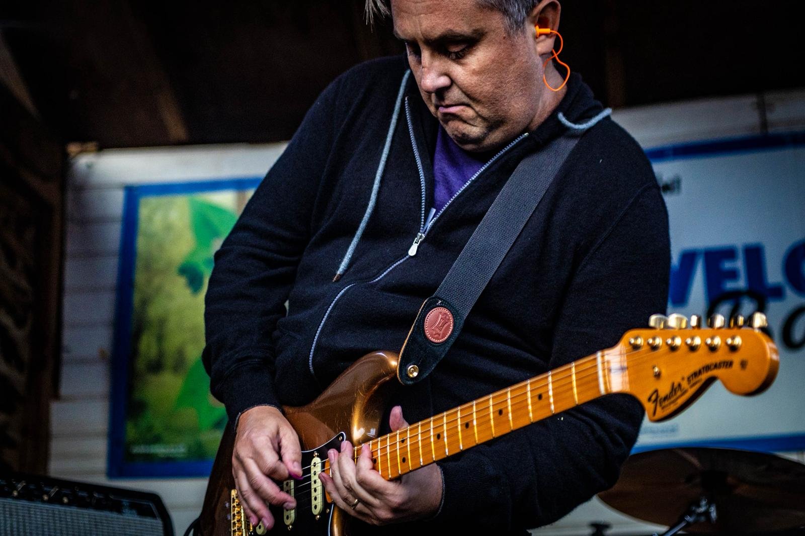 A person wearing a black hoodie and purple shirt, playing a brown Fender Stratocaster electric guitar with a black strap at an indoor event.