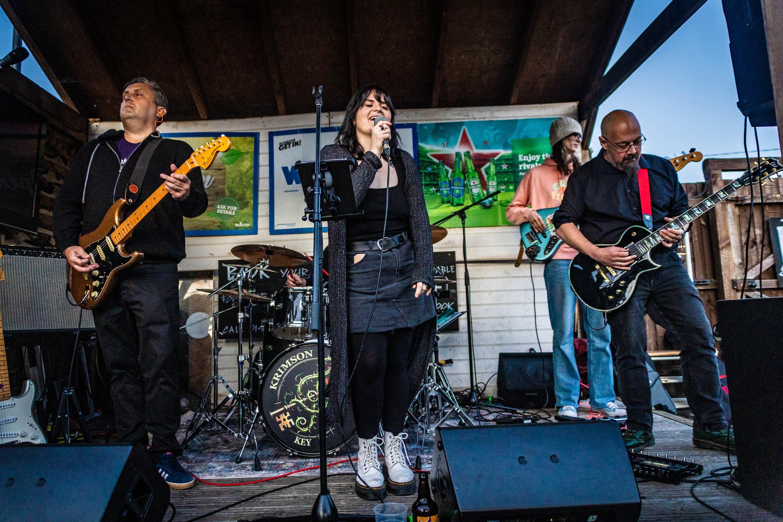 Band performing on stage with a male guitarist, a female singer, a male bass player, and a male guitarist. The stage has advertisements in the background, and they are under a wooden roof.