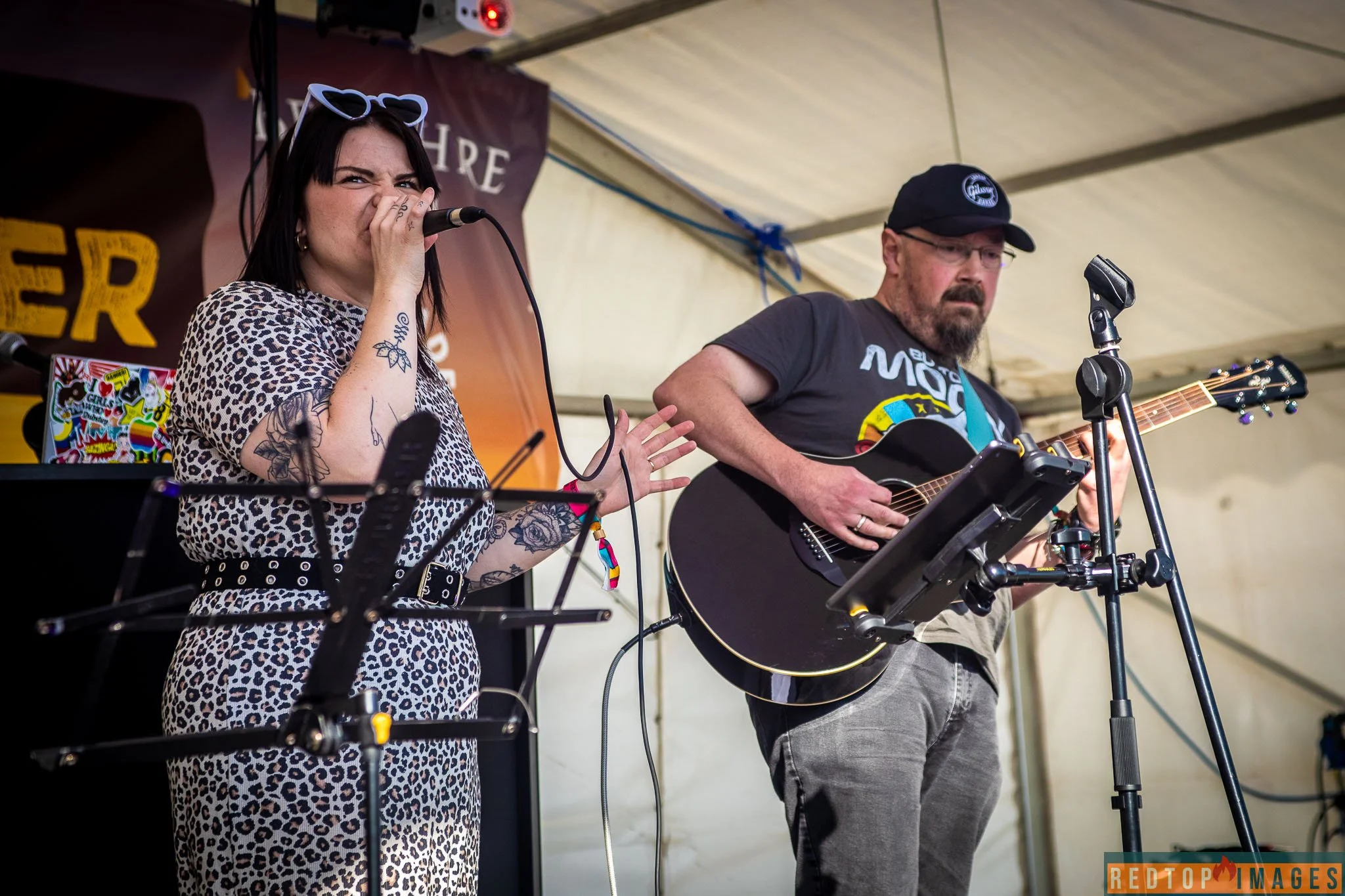 A woman with tattoos on her arm singing into a microphone and a man with a beard playing an acoustic guitar on stage under a tent.