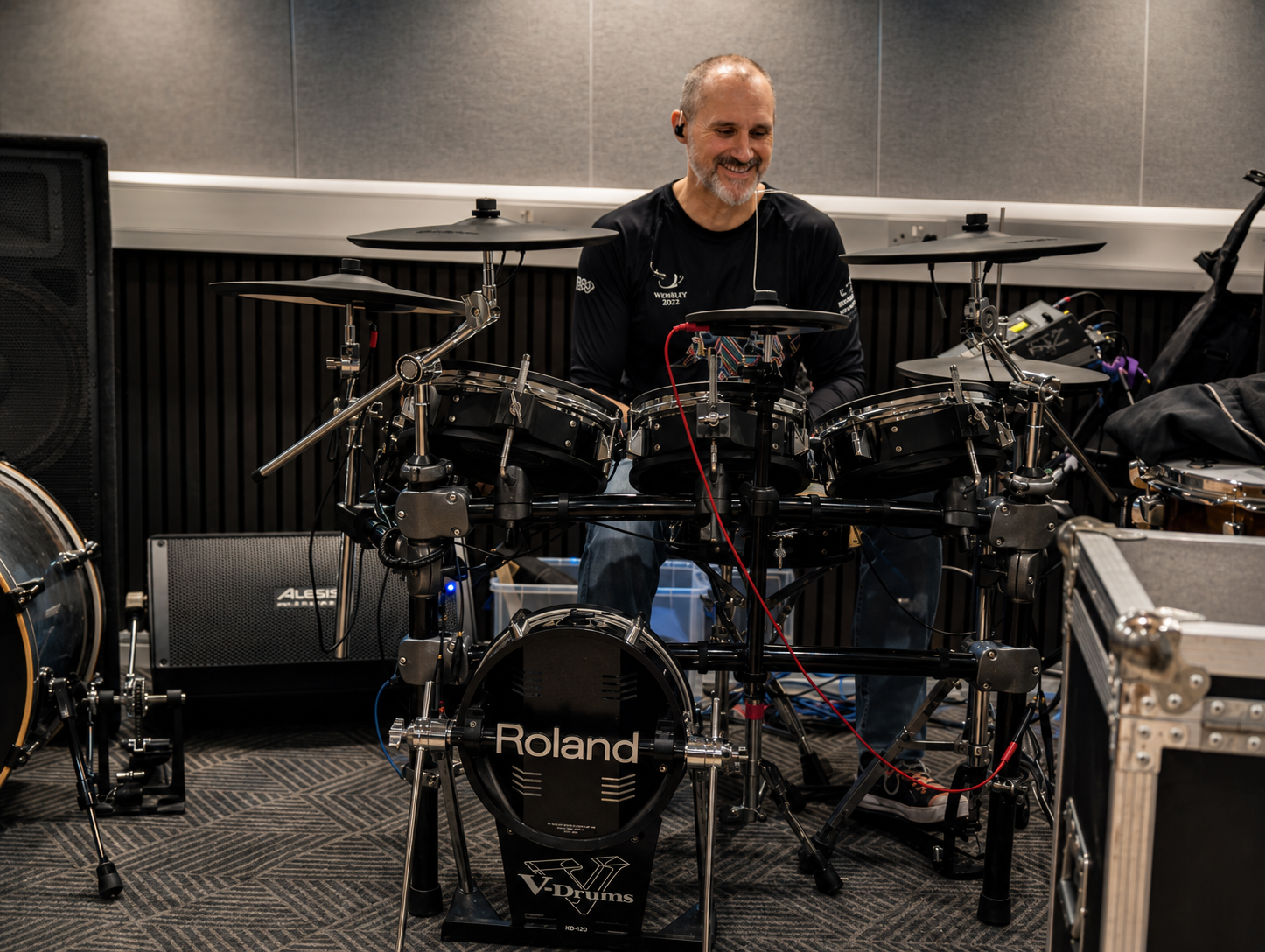 A man playing a digital drum set in a music rehearsal space, smiling and wearing a black shirt.