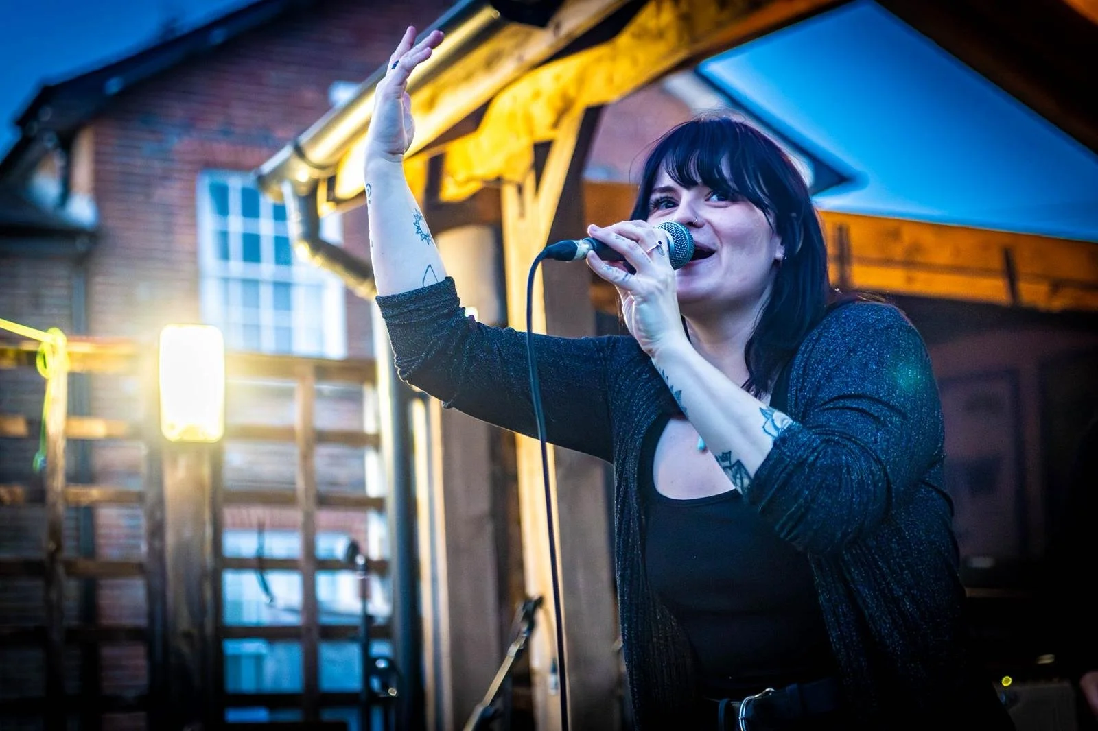 A woman singing into a microphone at an outdoor gathering during dusk, with a wooden structure and lights in the background.