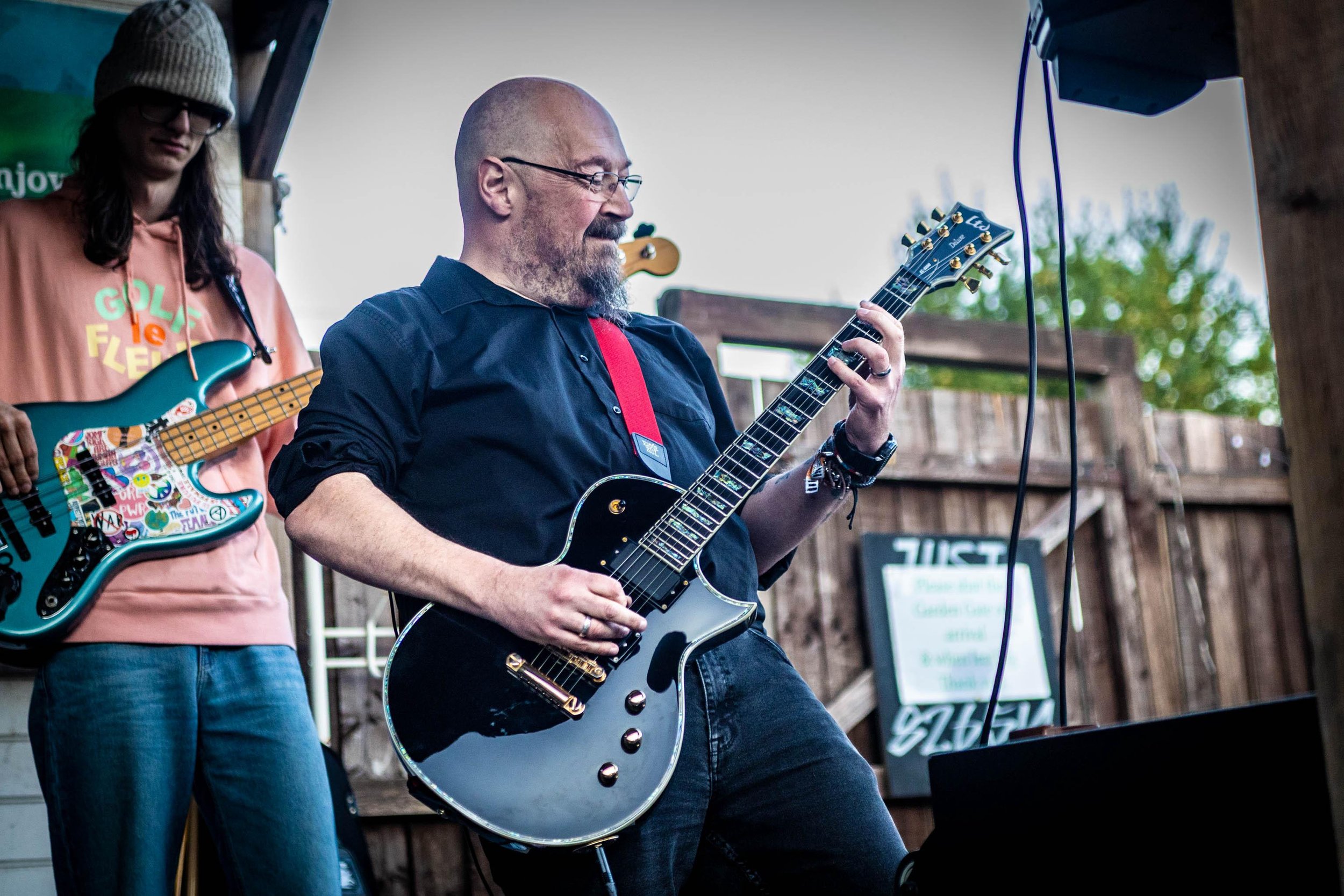 A man playing an electric guitar outdoors during a live music performance, with a Luky standing beside him holding another guitar, both of them are focused on their instruments.