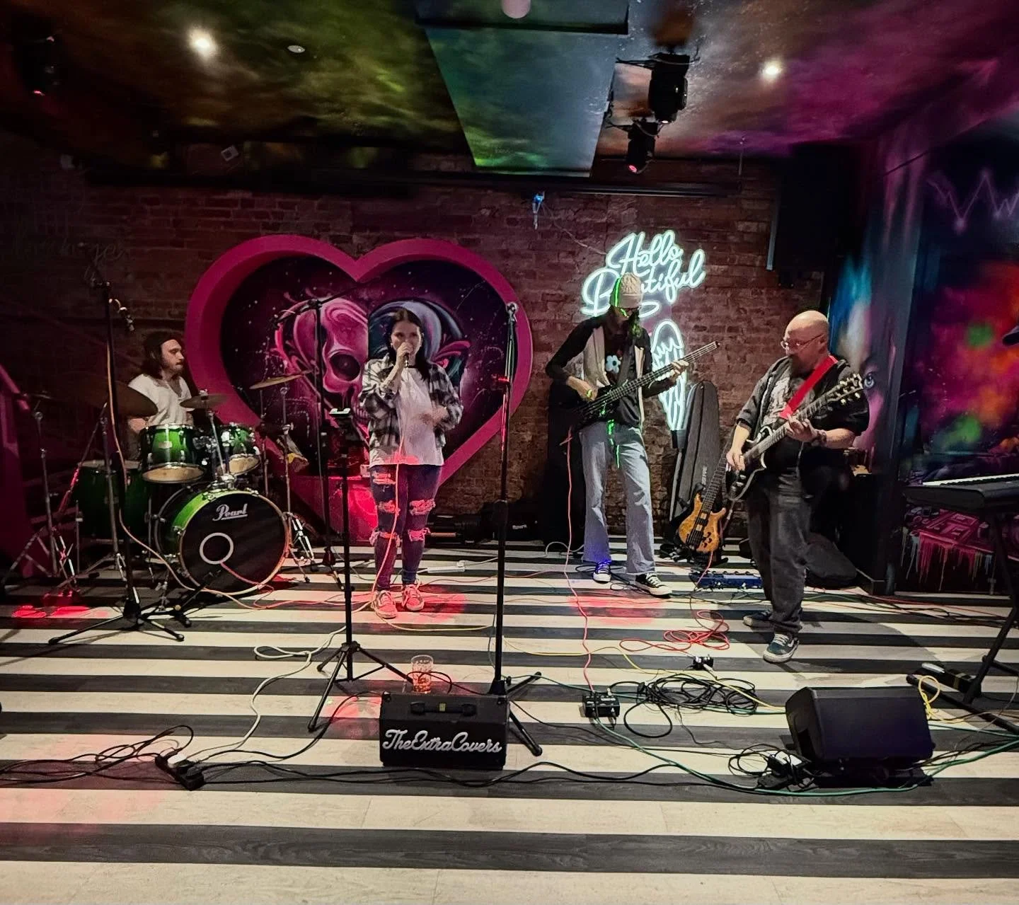 A band performing live on stage in a cozy venue with brick walls and colorful lights. The band includes a drummer, a female singer, and two guitarists. The backdrop features a pink heart with a skull and a neon sign that reads 'Hello Beautiful.'