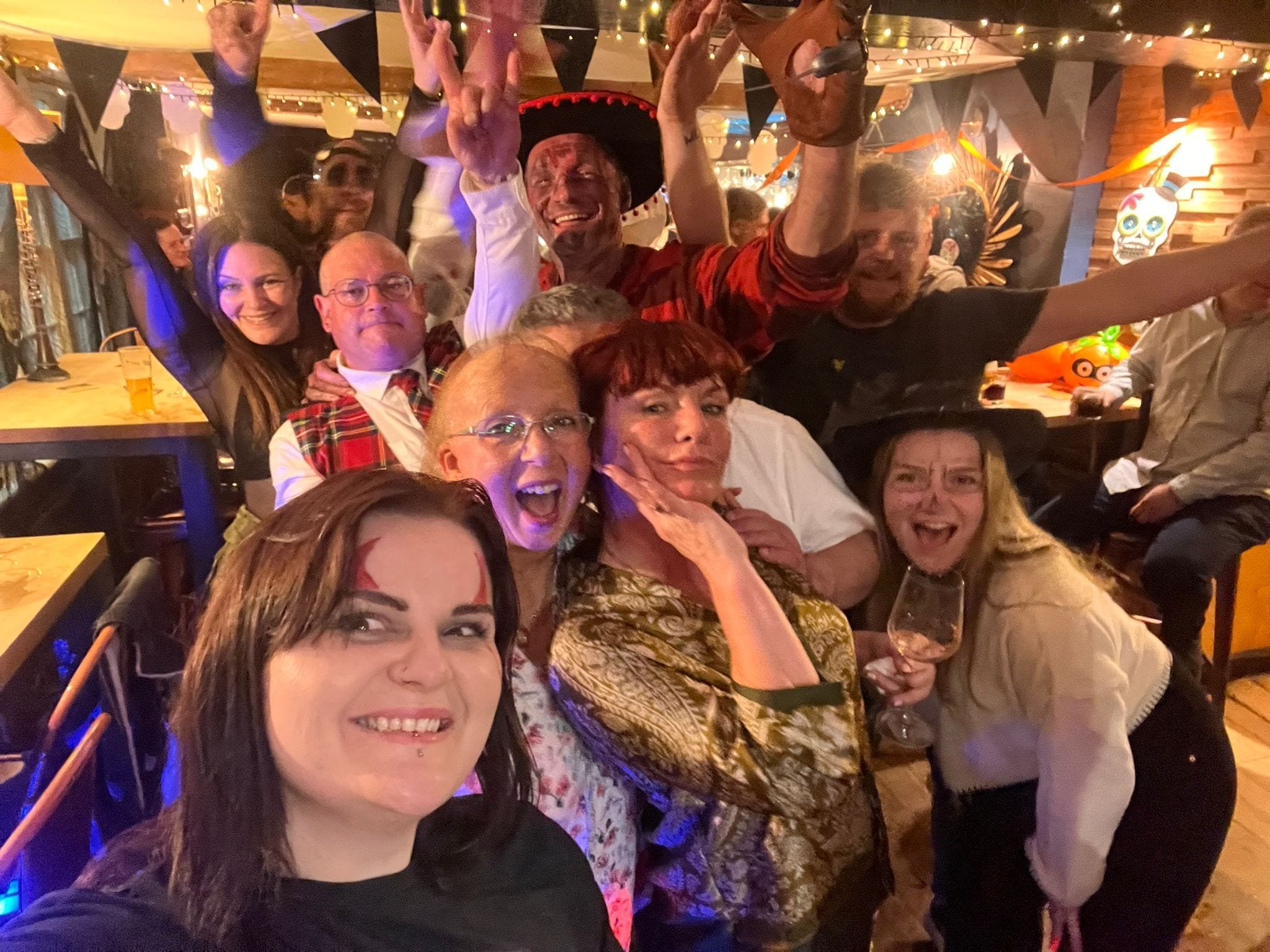 Group of people celebrating at a festive party, with some wearing costumes and headpieces, and decorations in the background including skull masks and string lights.