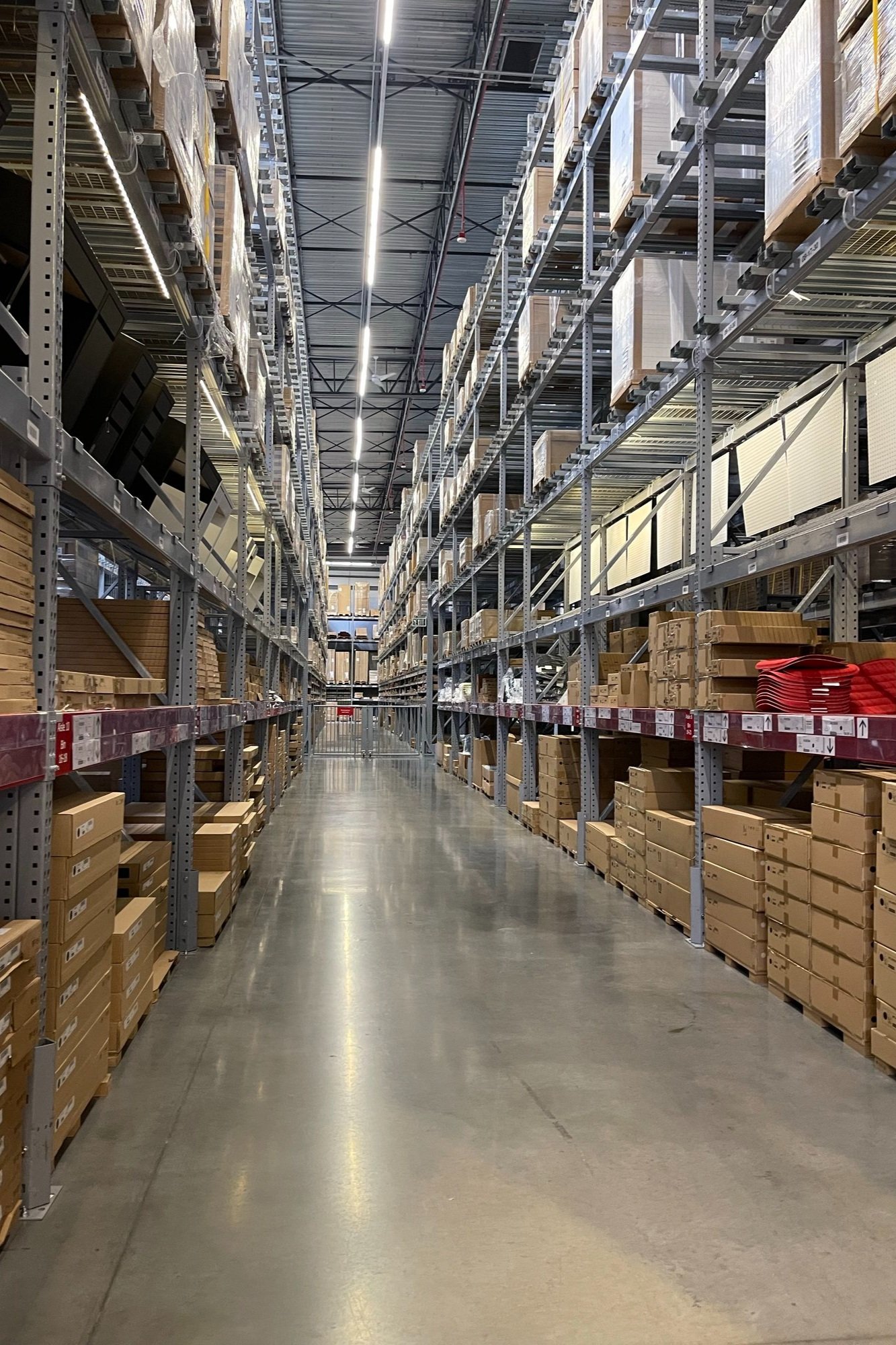 Aisle in a warehouse with high metal shelving filled with boxes and products, illuminated by overhead fluorescent lights.