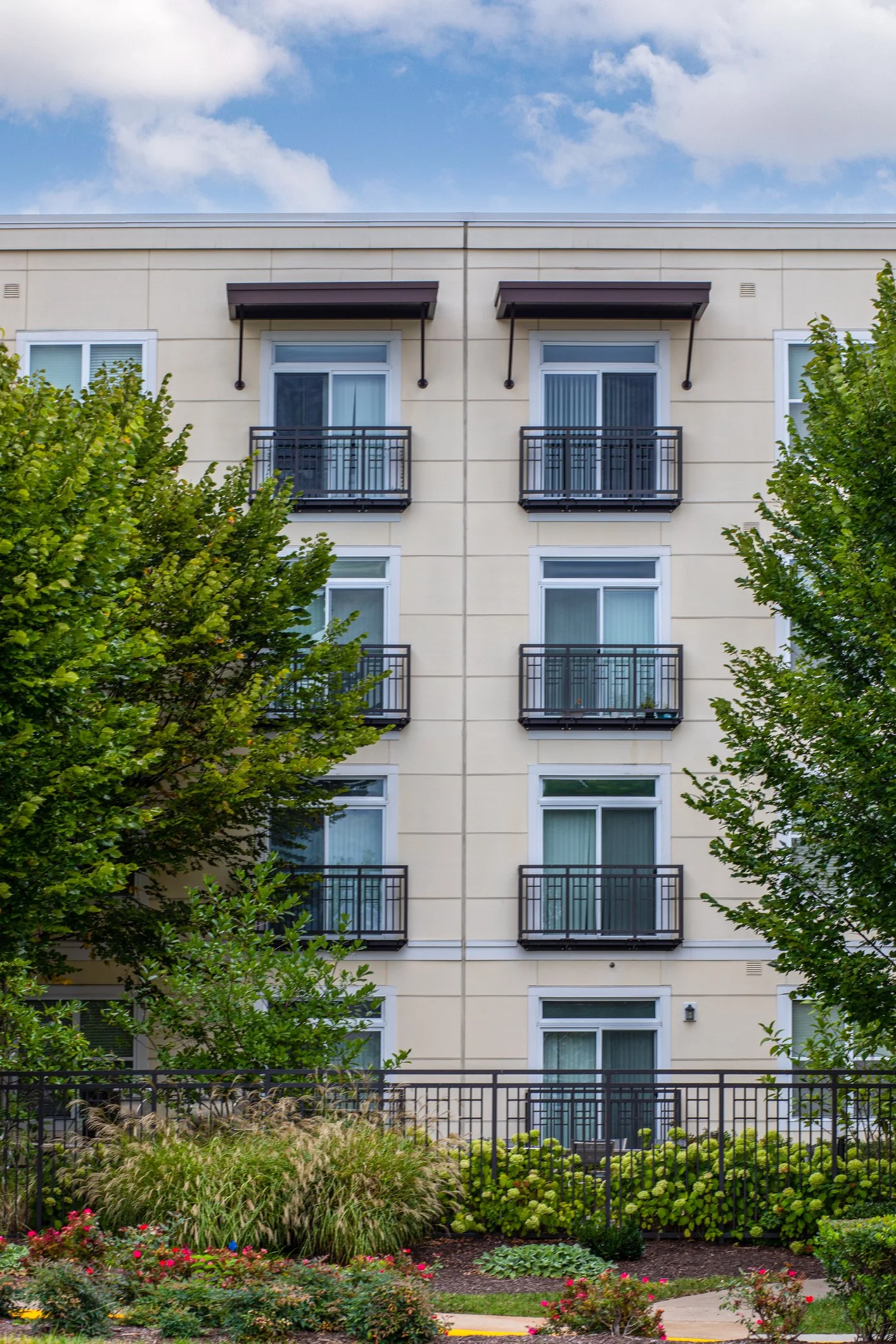 The image shows a beige apartment building with four floors, balconies on each level, and two large trees partially obstructing the view. The sky is partly cloudy with blue patches visible, and there is a landscaped garden with flowering plants, bushes, and ornamental grasses in the foreground.