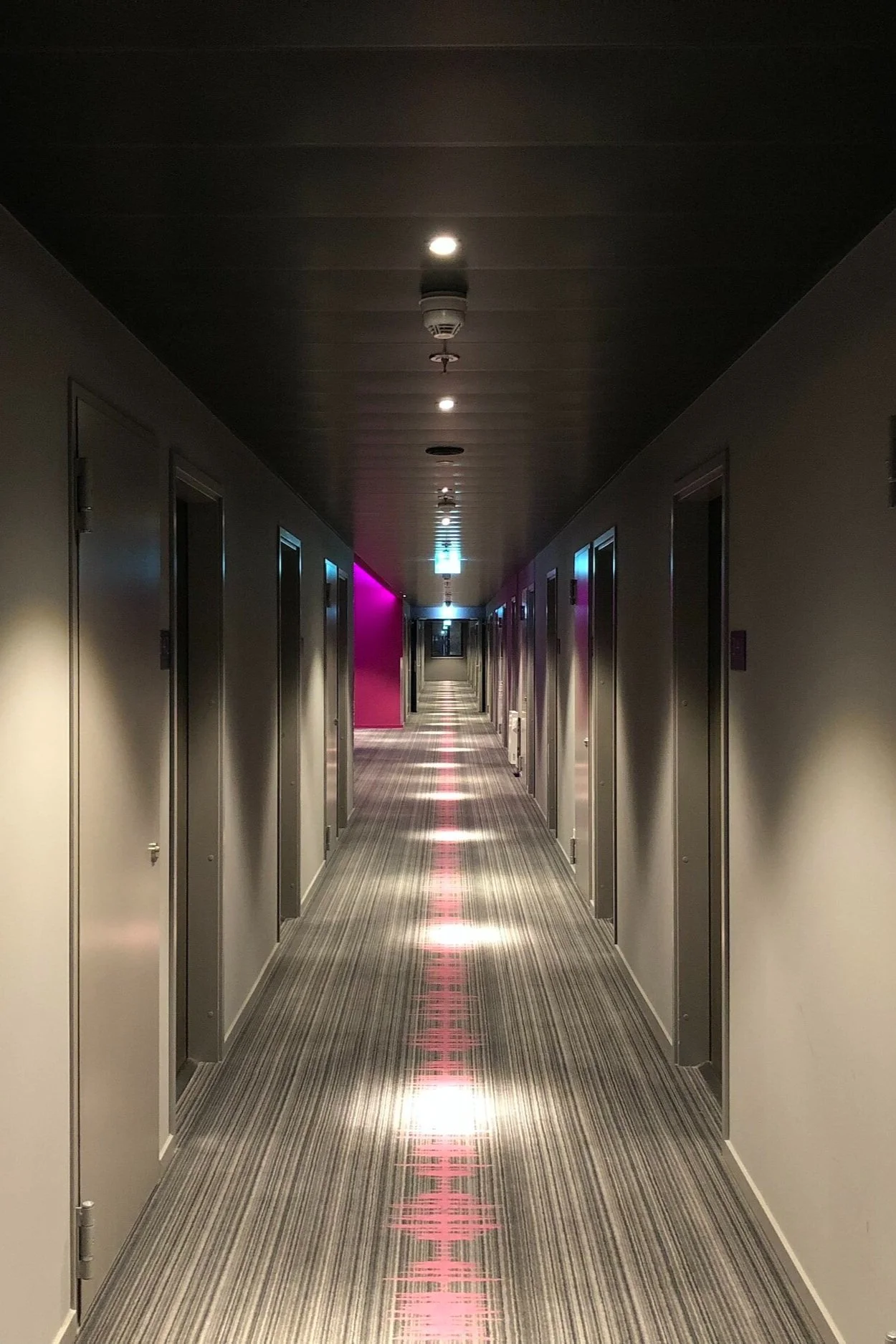 Empty hotel corridor with closed doors on both sides, patterned carpet, and colorful lighting on the walls, with some ceiling lights turned on.