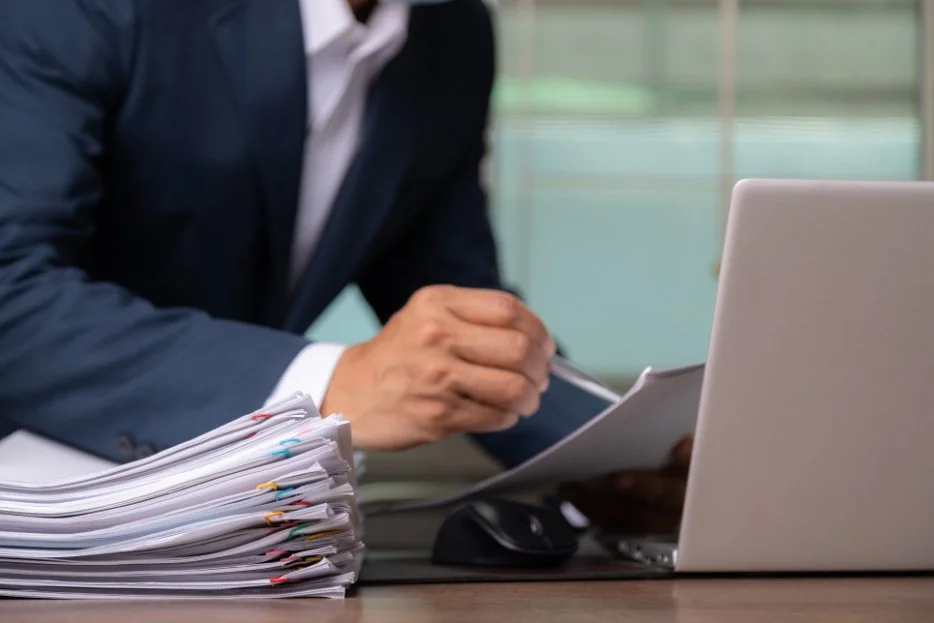 A person in a business suit sitting at a desk with a large stack of documents, a laptop, a computer mouse, and holding some papers, in an office environment.