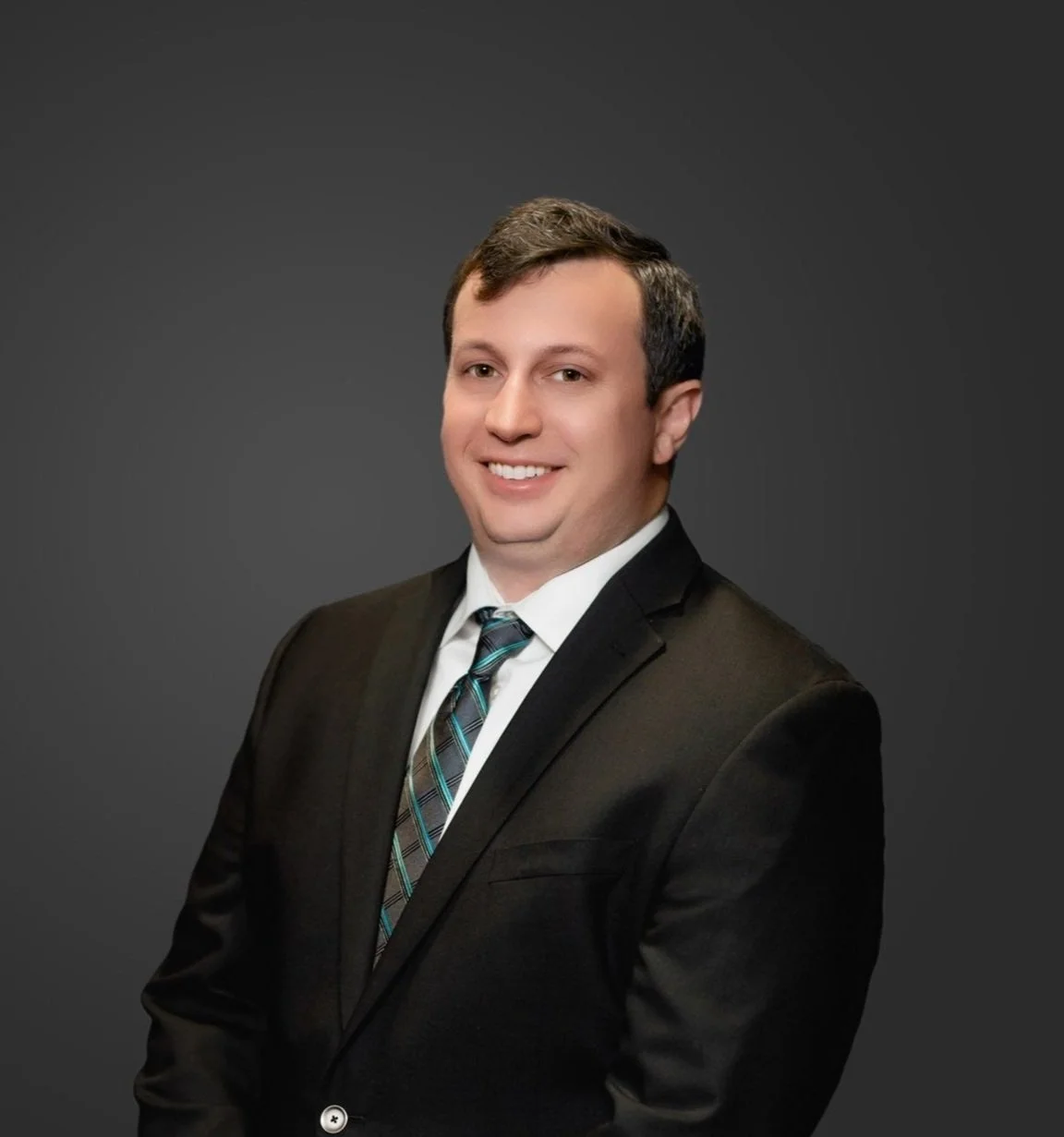 Professional headshot of a man in a black suit, white shirt, and striped tie, smiling against a dark gray background.