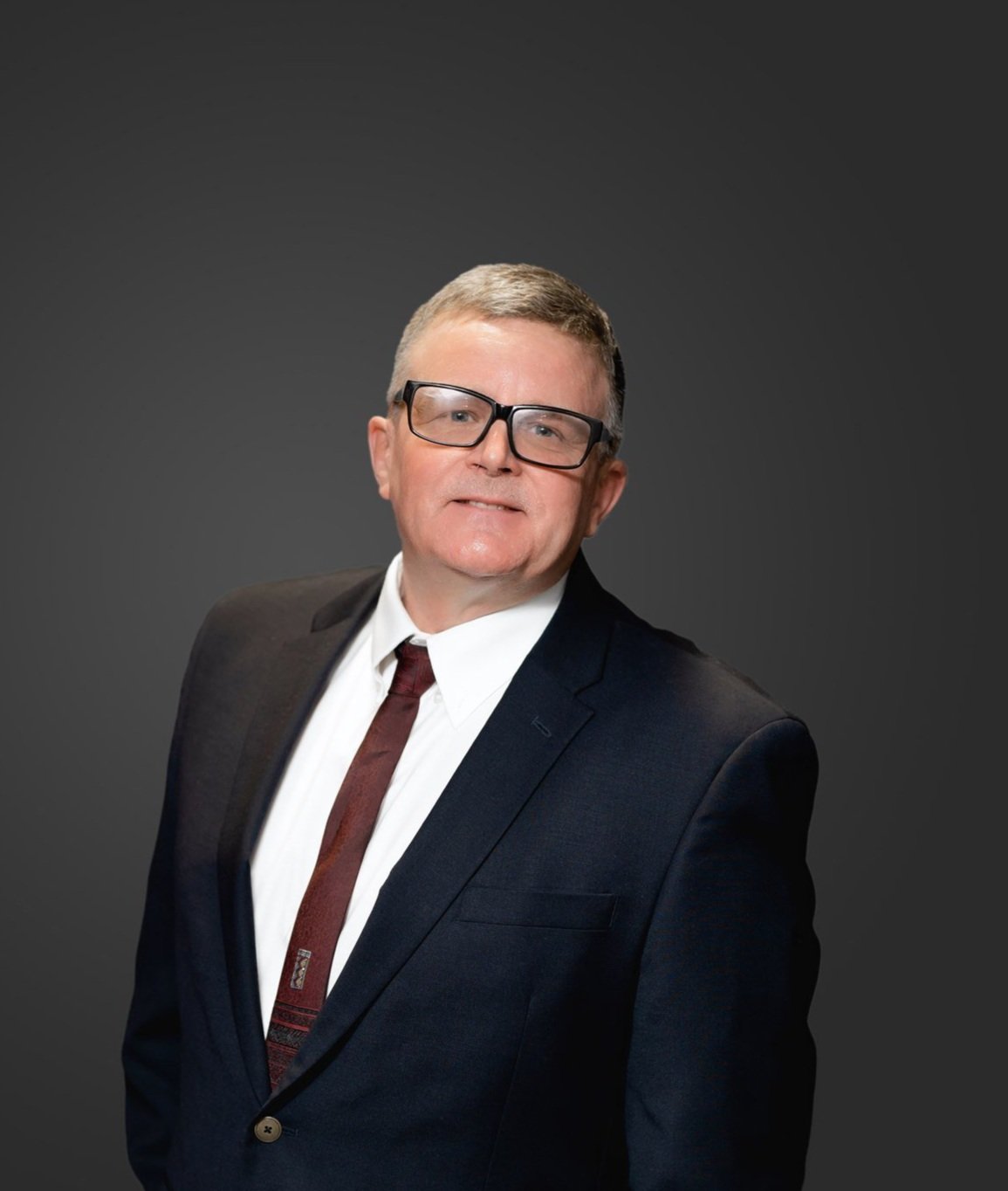 A professionally dressed man in a dark suit, white shirt, and maroon tie standing against a dark gray background.