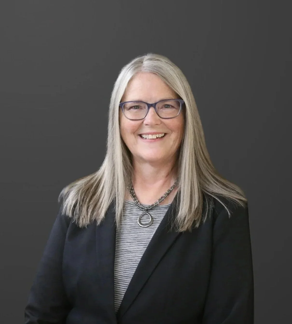 A smiling woman with long gray hair, wearing glasses, a black blazer, a striped shirt, and a silver necklace with a round pendant, standing against a plain dark background.